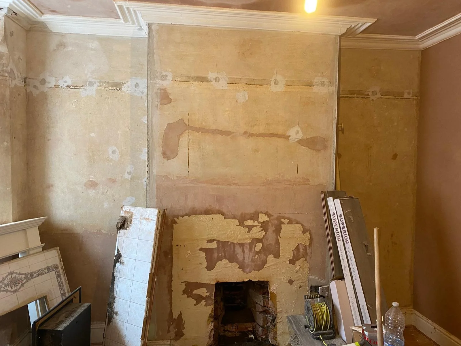 A room undergoing renovation with peeling paint, exposed drywall, and construction tools like a yellow extension cord, a bottle of water, and a stack of flooring material.