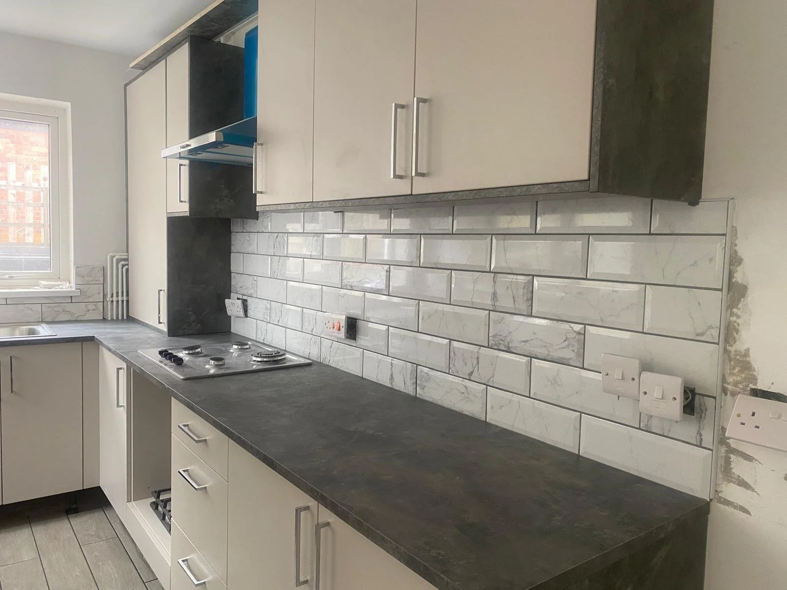 Modern kitchen with grey countertop, white cabinets, marble tile backsplash, and a gas stovetop under a window.