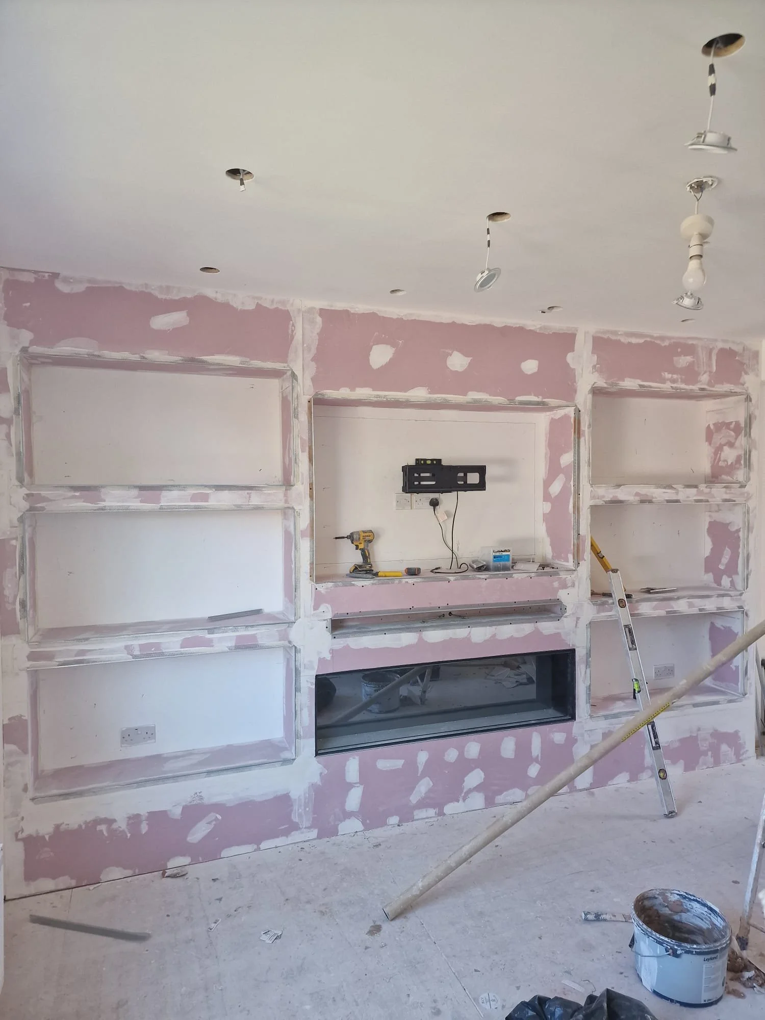 Living room under renovation with unfinished wall paneling, open shelving, and installation of lighting fixtures in progress.