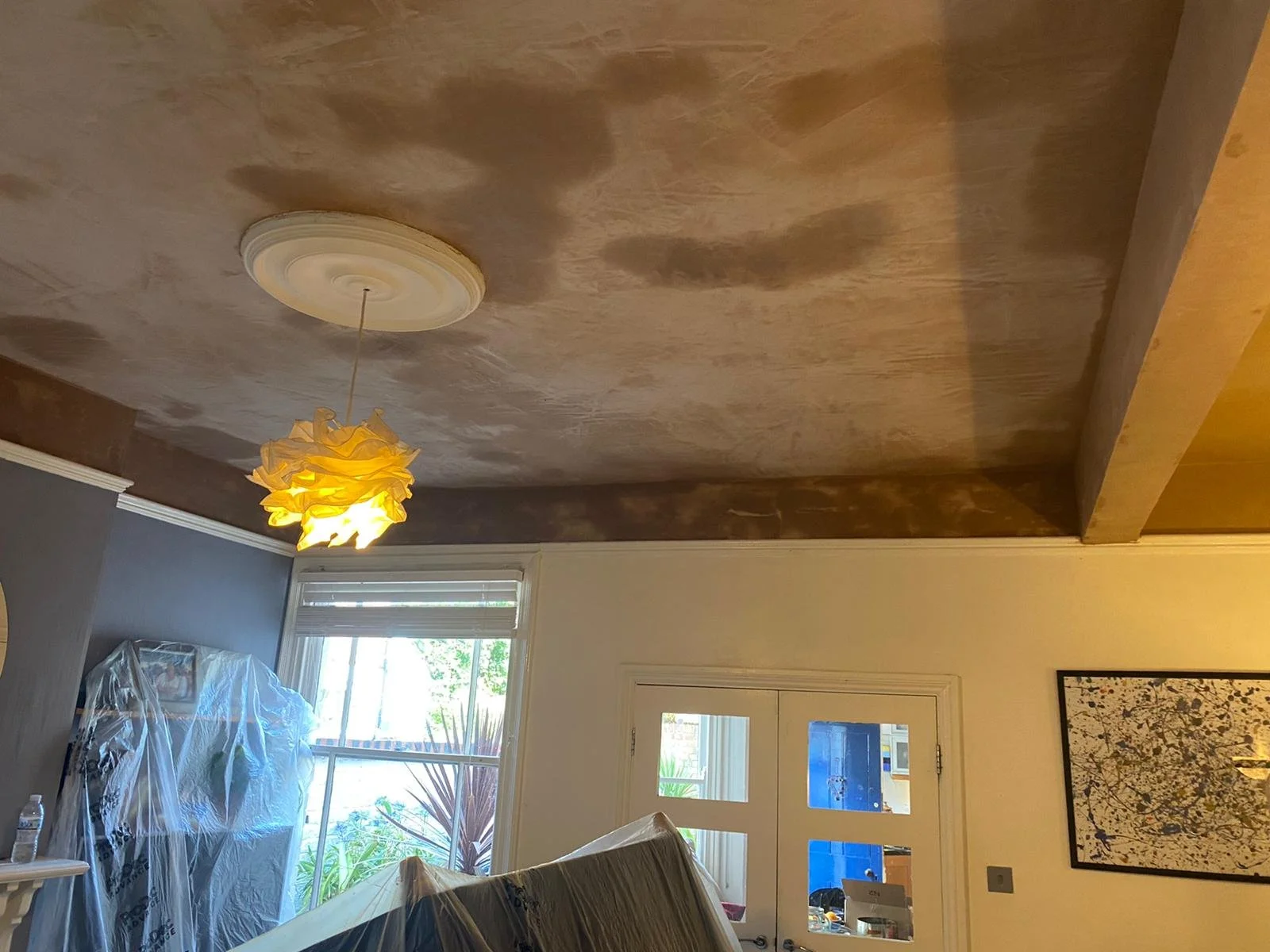 Living room with a ceiling light fixture, large window, and a door, with some furniture and artwork on the walls, and a couch covered with plastic.