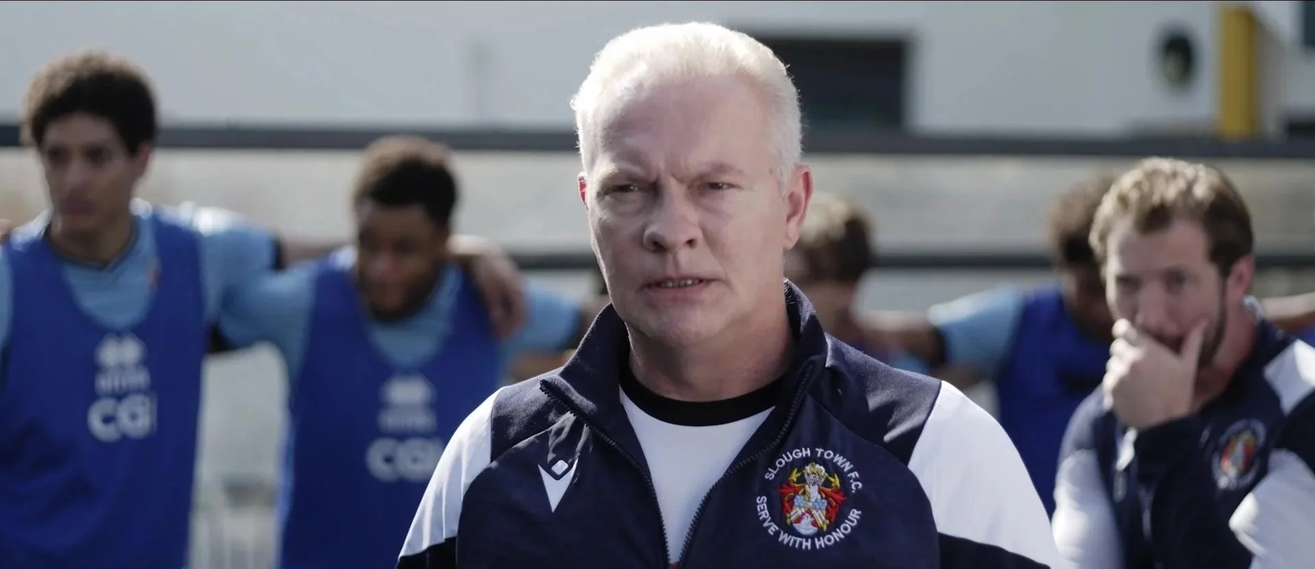 A man with white hair, wearing a blue sports jacket with a 'Slough Town FC' emblem, appears to be coaching or giving instructions to a group of young soccer players standing behind him with arms around each other's shoulders, on a soccer field.
