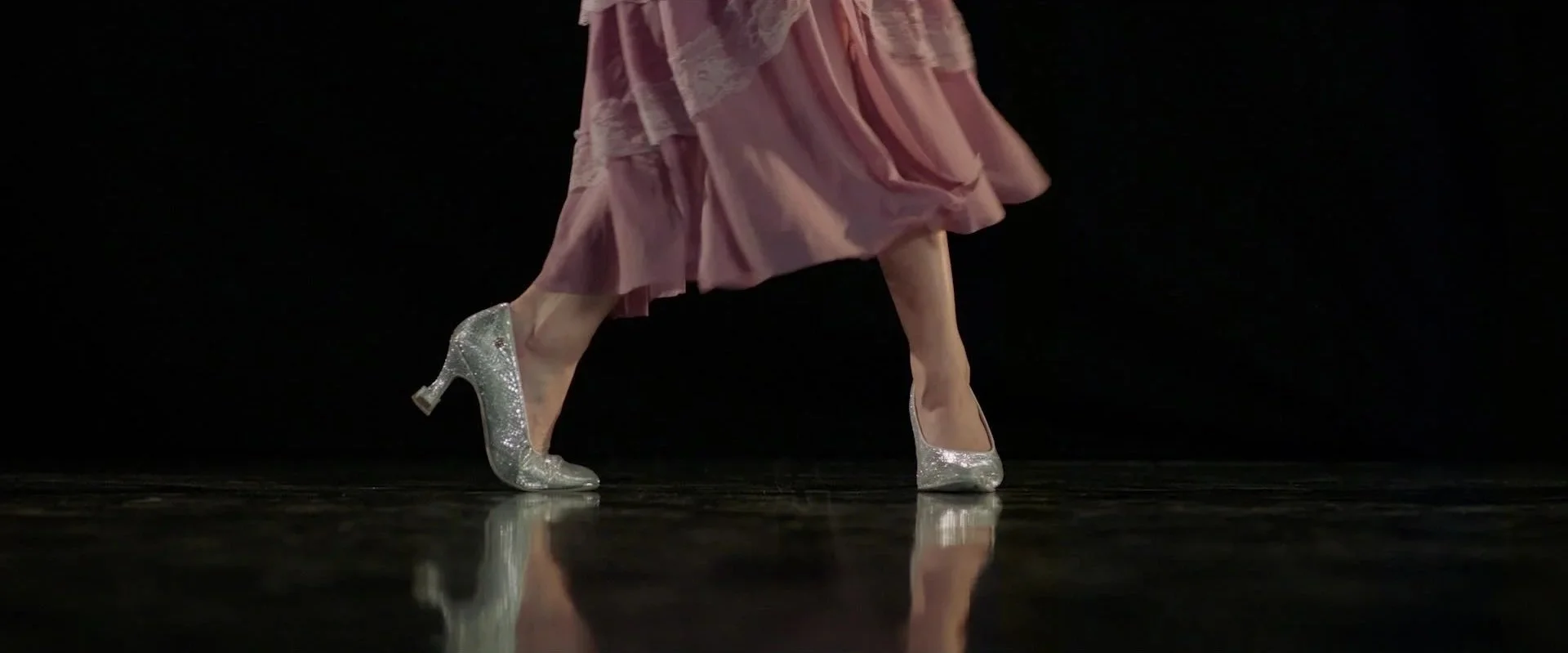 Close-up of a dancer's legs and feet on stage; she is wearing a pink skirt and sparkling silver high-heeled shoes.