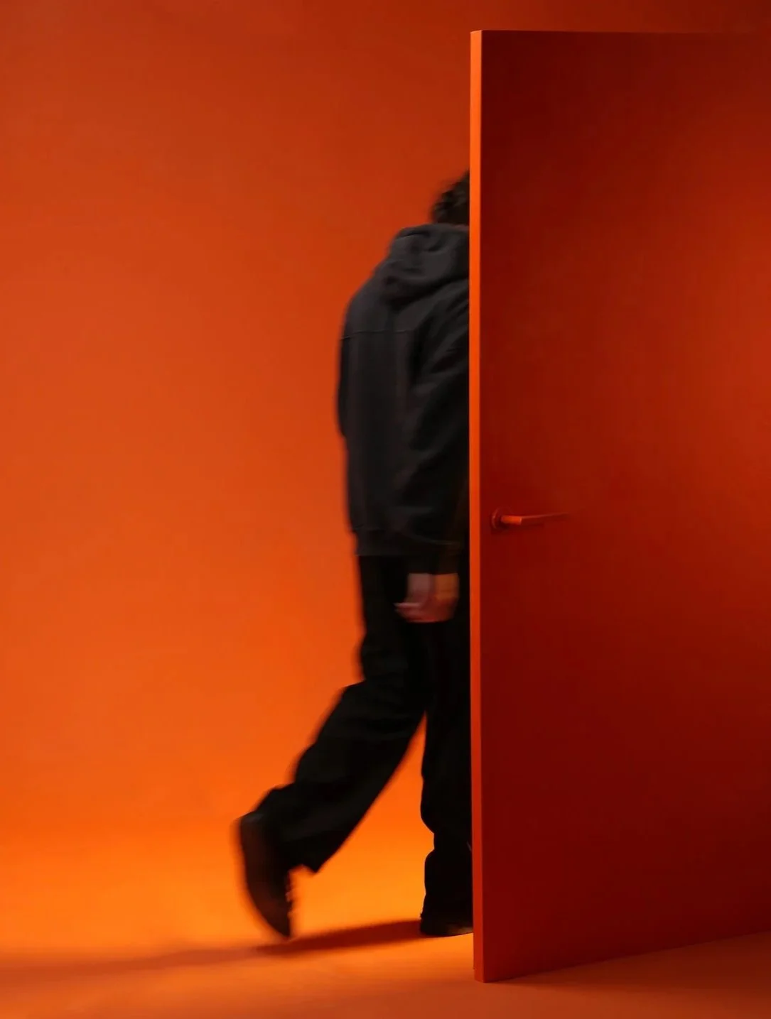 A person dressed in black walking out of an orange-colored door against an orange background.