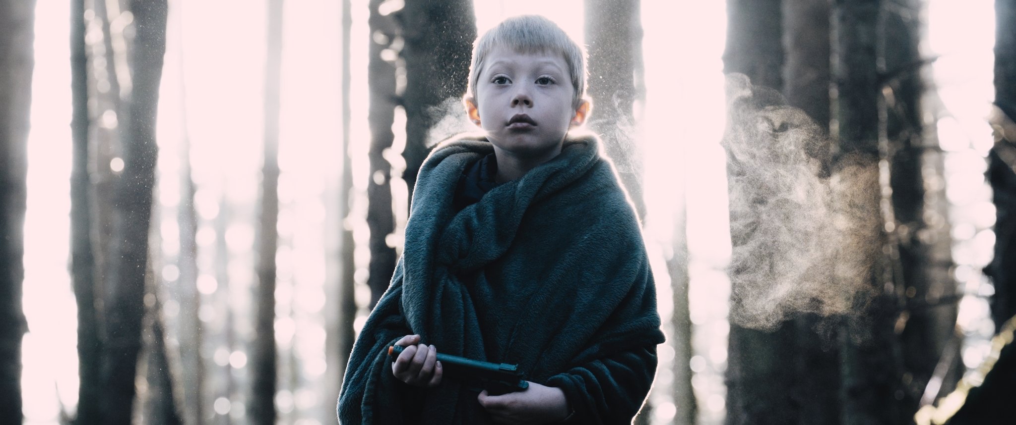 A young boy with short hair and a serious expression, wearing a dark hoodie and wrapped in a blanket, standing in a foggy forest with tall trees and sunlight streaming through the branches.