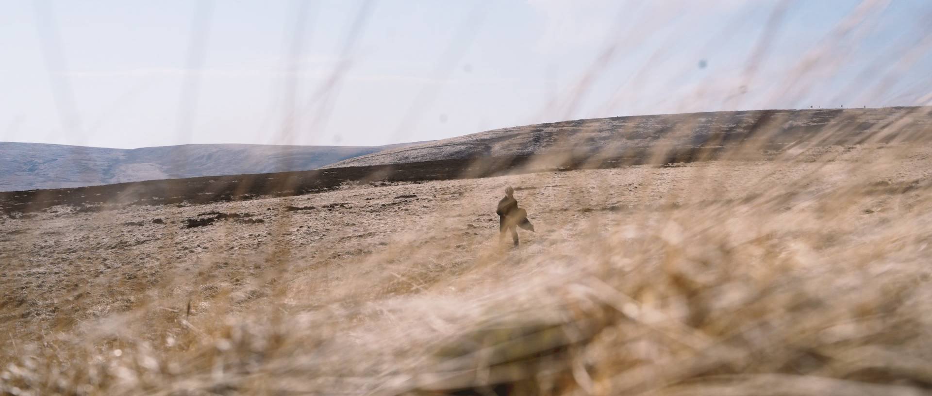 A person in a brown jacket walking through a dry, grassy landscape with a hill in the distance under a cloudy sky.