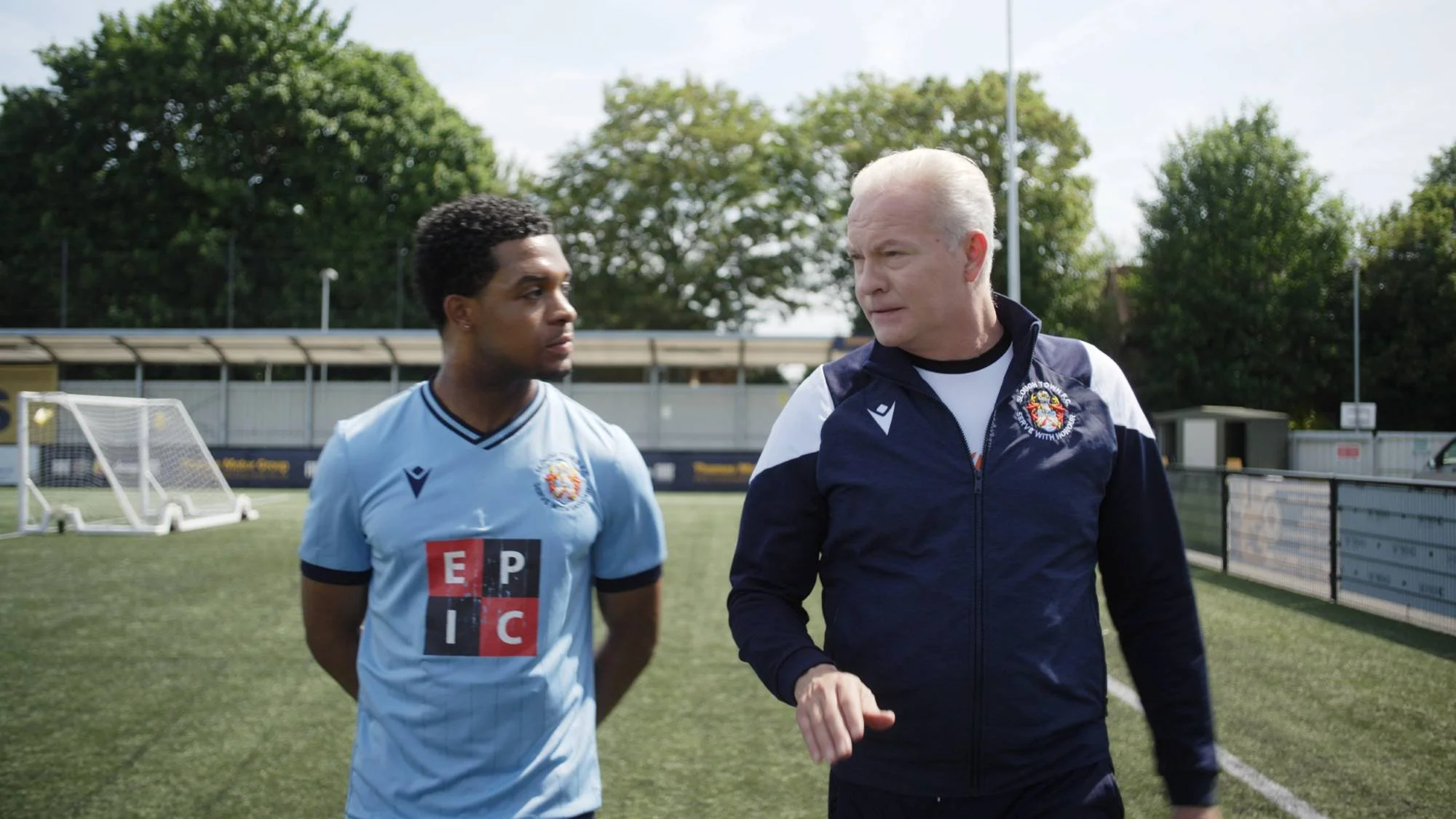 Two men walking on a soccer field, one in a light blue soccer uniform and the other in a navy tracksuit, with trees in the background.