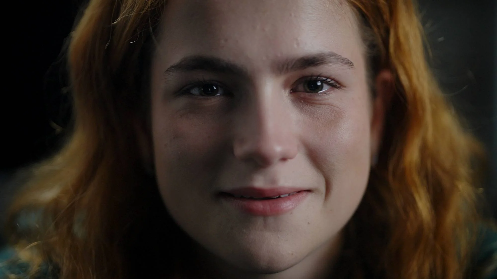 Close-up of a young woman with red hair, green eyes, and light skin, looking directly at the camera with a slight smile.