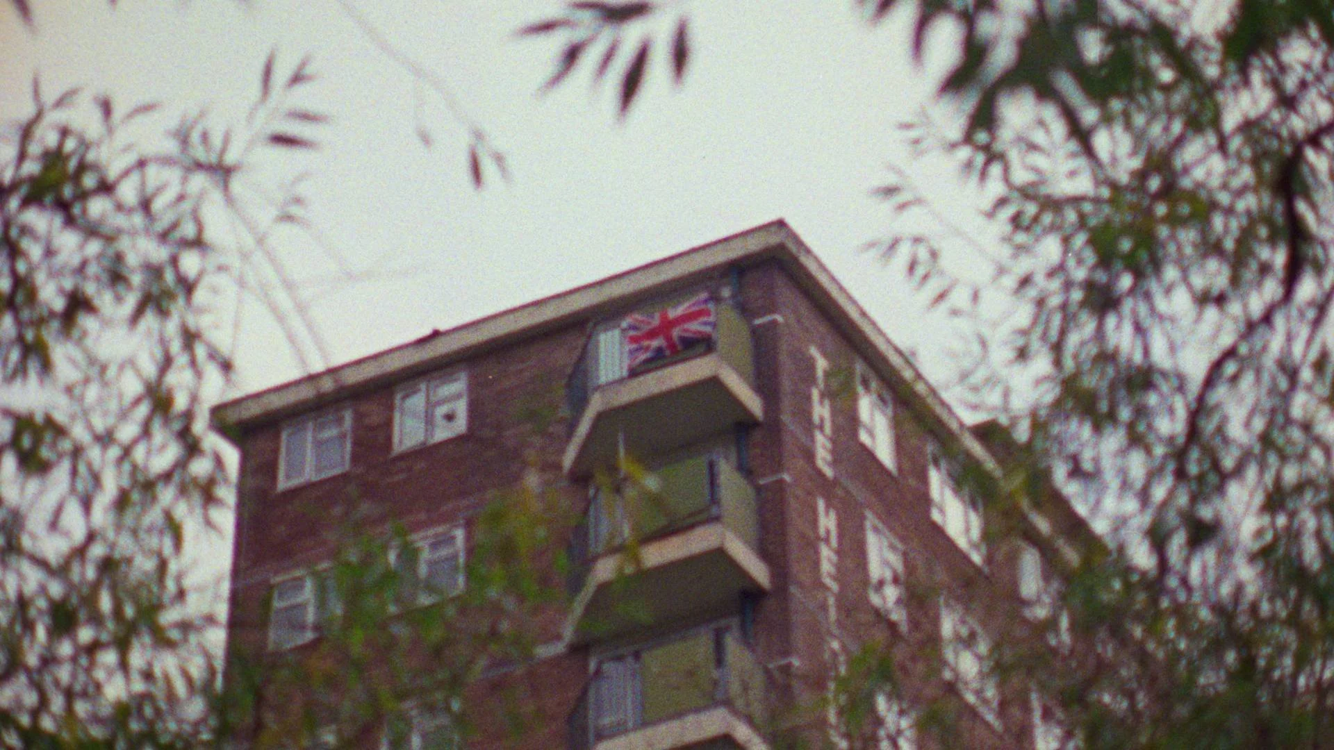 A multi-story brick building with balconies, displaying a Union Jack flag and the words 'THE HOTEL' on the side, surrounded by tree branches.