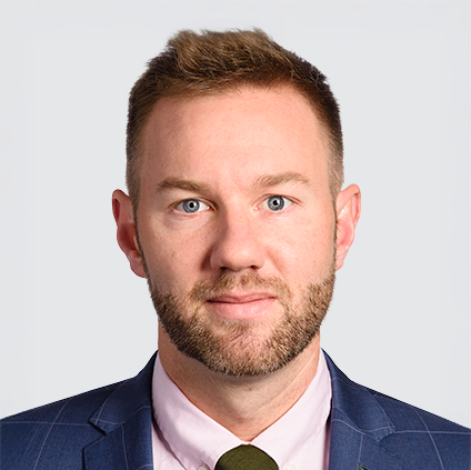 Headshot of a man with short brown hair and a beard, wearing a navy blazer, light pink shirt, and dark tie, against a light gray background.