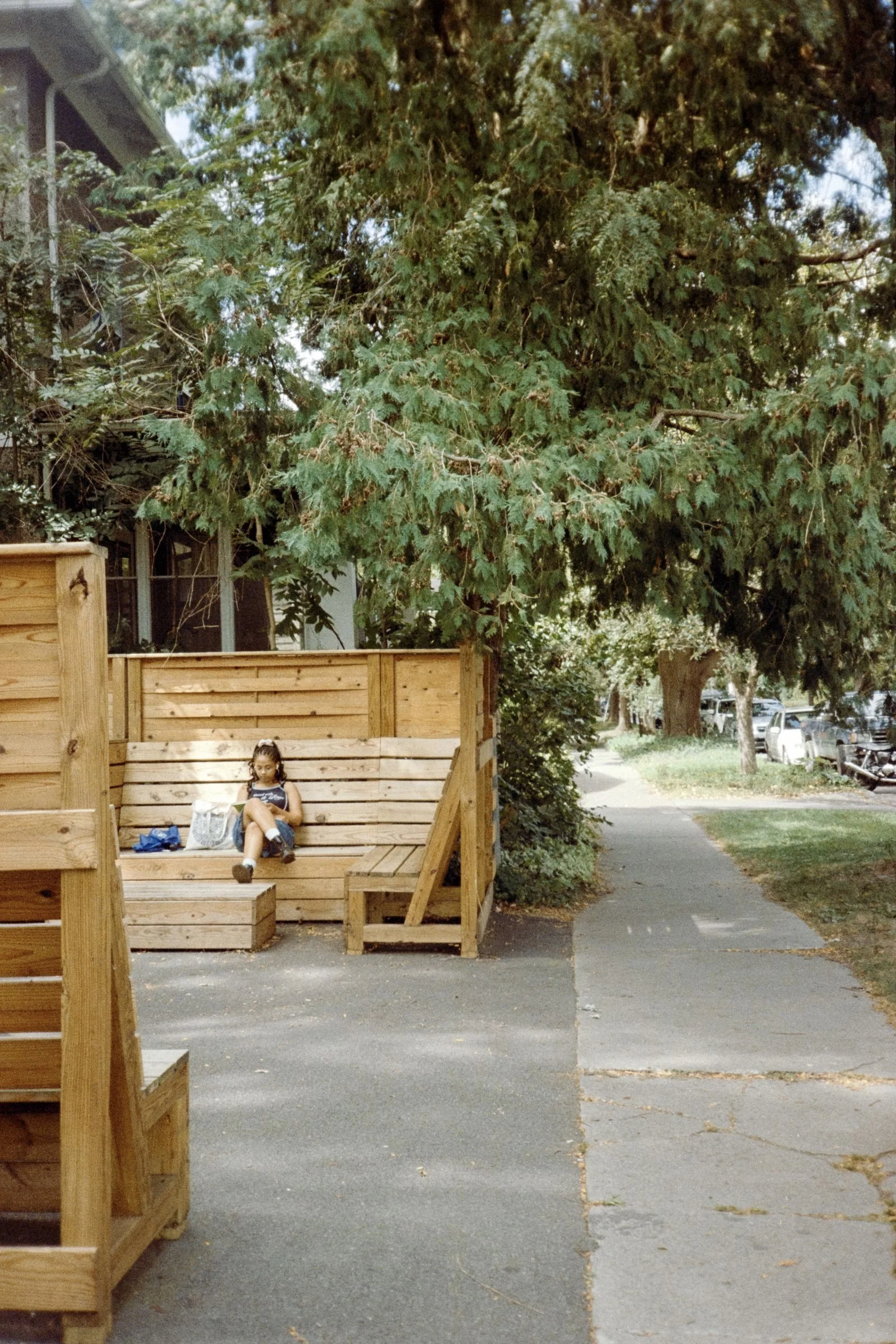 A young girl sitting on a wooden bench next to a wooden fence, reading a book with a bag beside her, on a sidewalk with trees in the background.