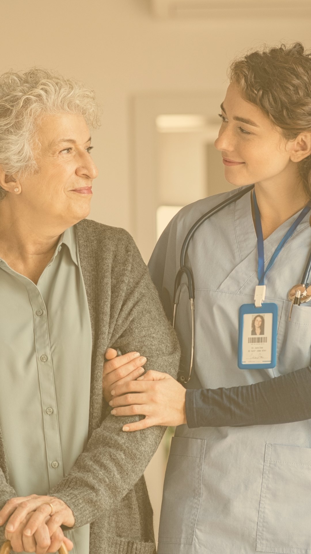 A healthcare professional, wearing scrubs and a stethoscope, gently holds the arm of an elderly woman who is smiling at her.