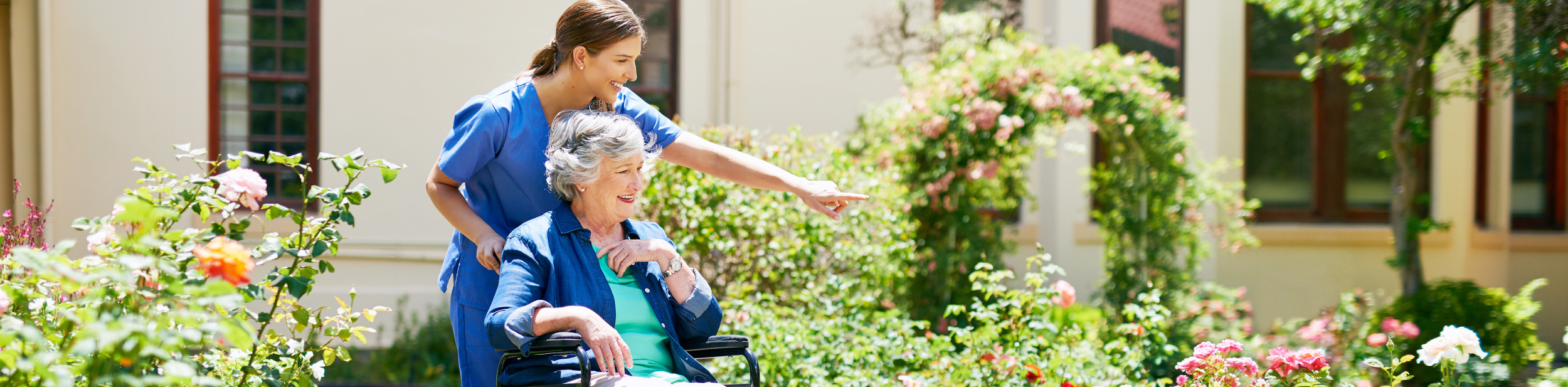 A young caregiver outdoors with an elderly woman in a wheelchair, smiling and pointing at flowers in a garden.