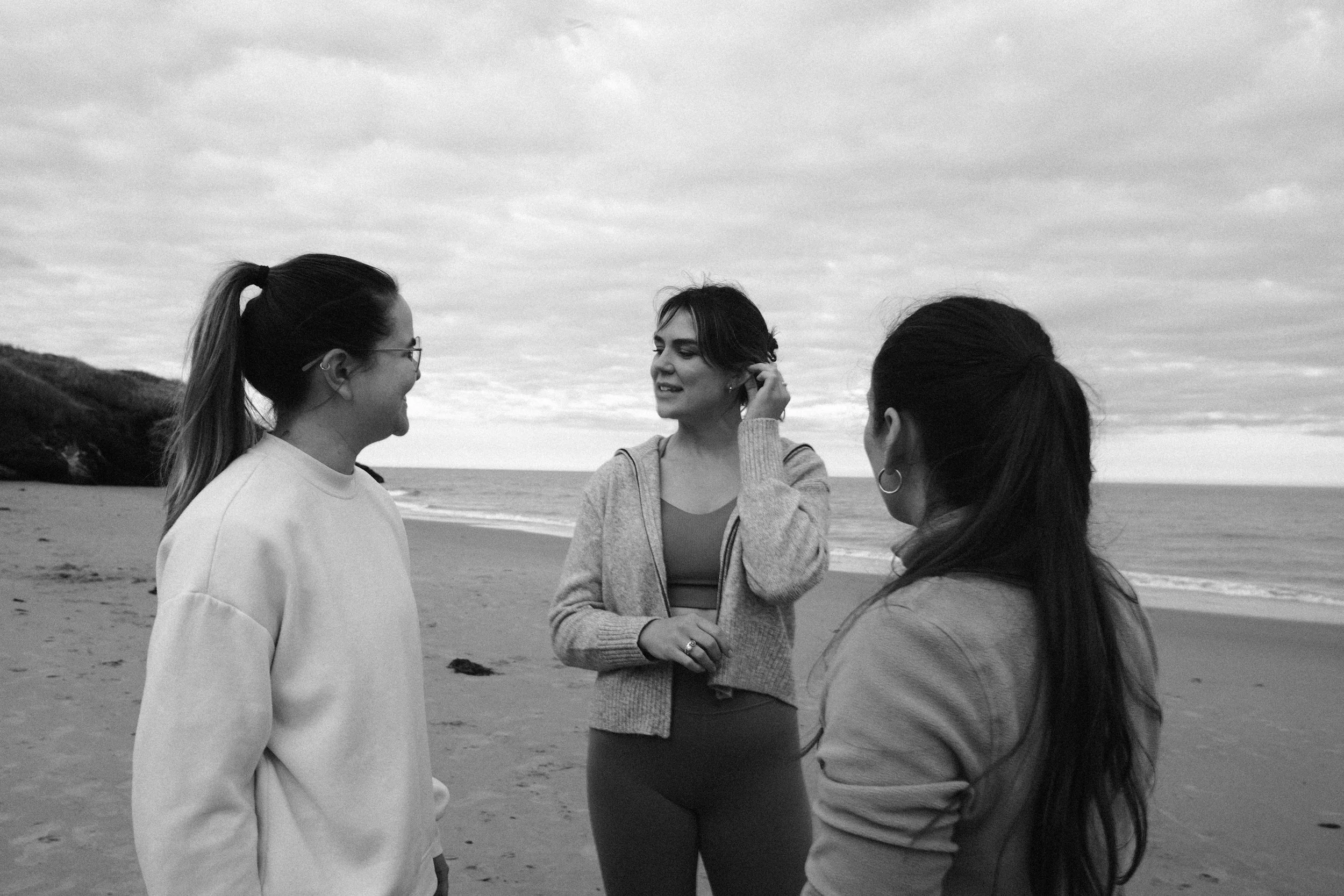 Four women standing on a beach, talking, with an overcast sky and a hillside in the background.