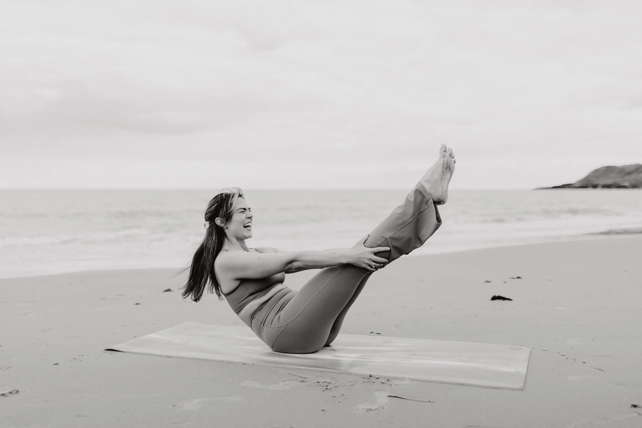 A woman practicing yoga on a beach, sitting in a boat pose with her arms wrapped around her legs, smiling and looking joyful.