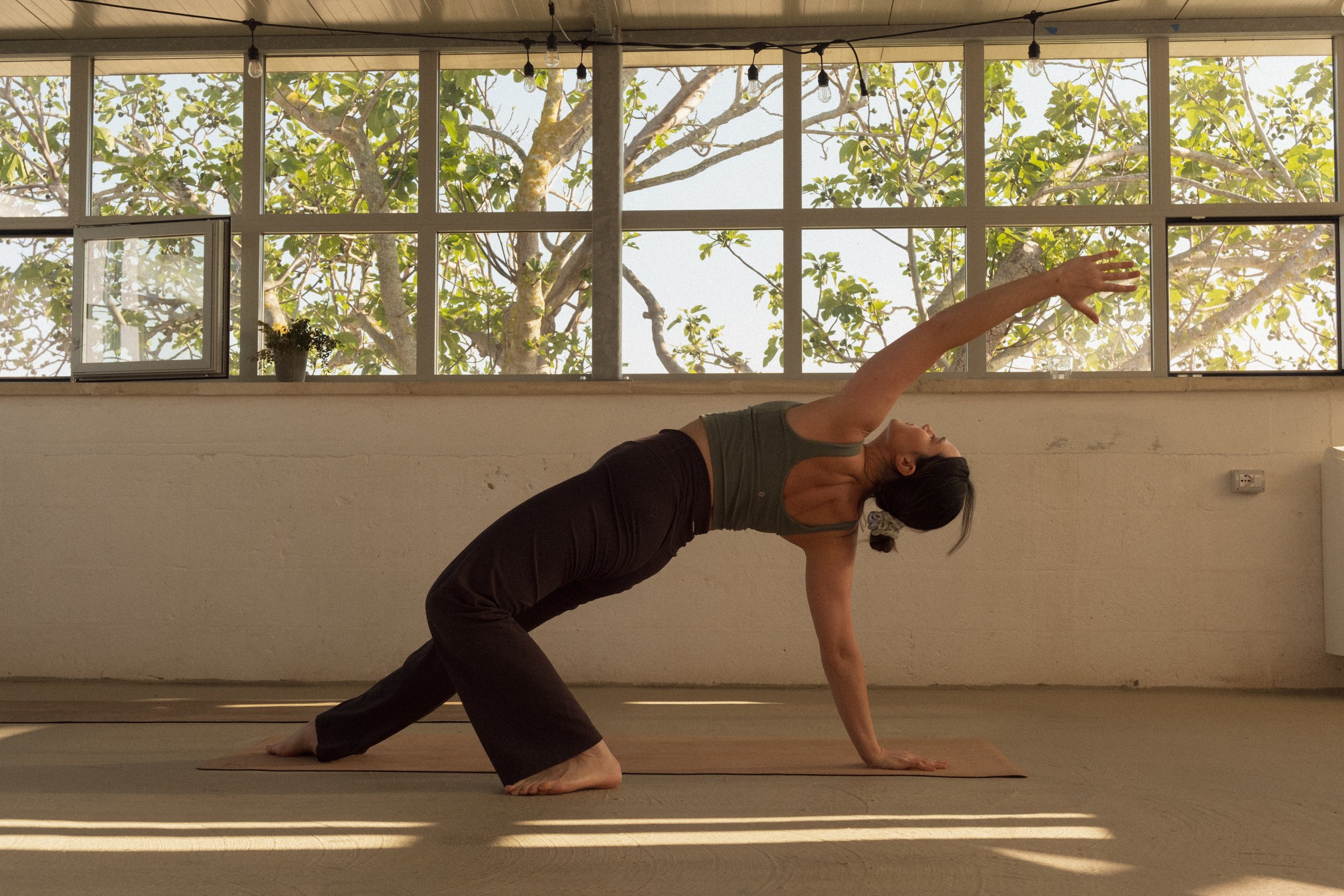 A woman practicing yoga indoors, in a pose resembling a modified downward dog with one arm extended forward, with large windows and trees outside.