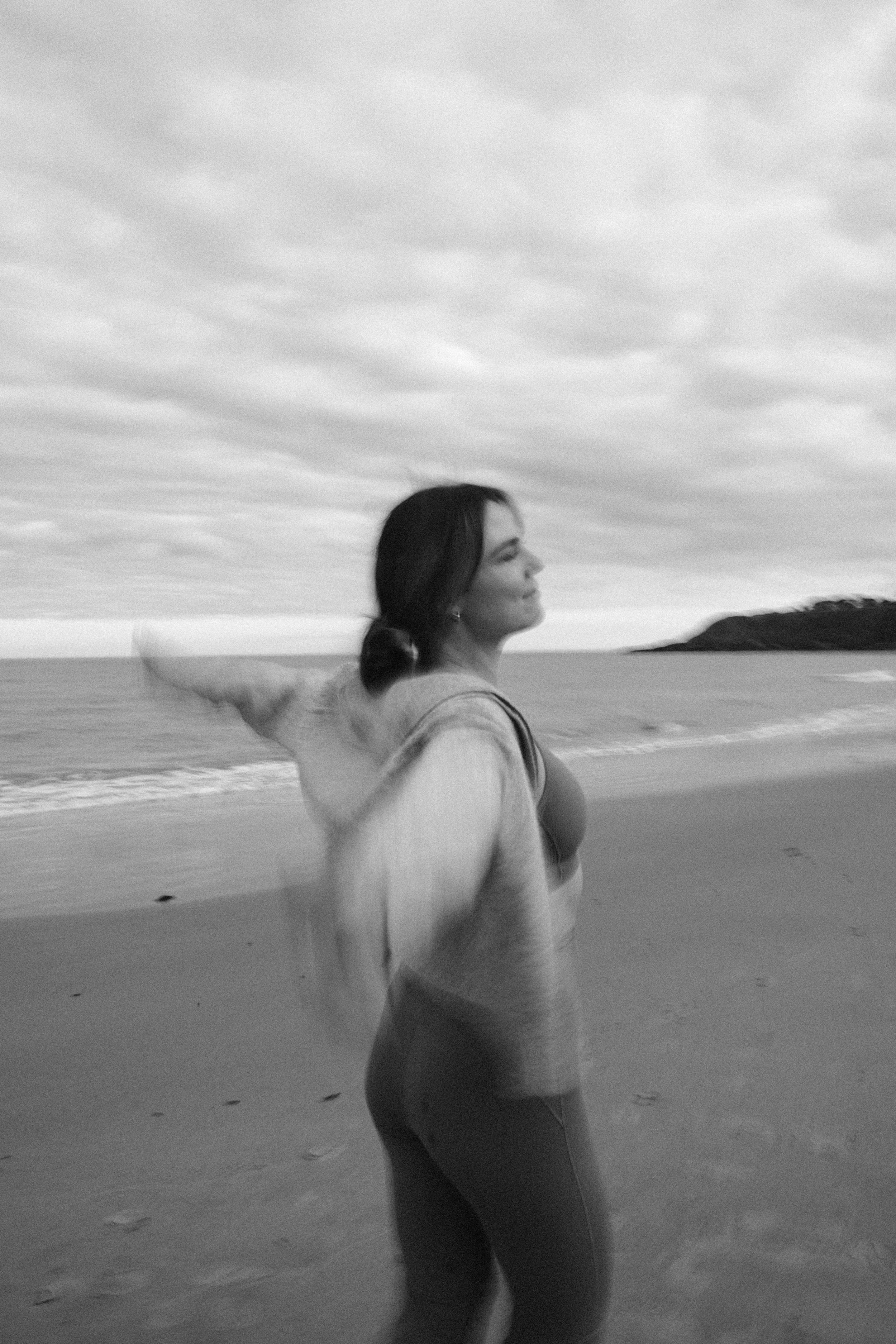A woman standing on a beach with her eyes closed and face turned to the sky, wearing a tank top and leggings, with an ocean and a distant landmass under a cloudy sky in the background.