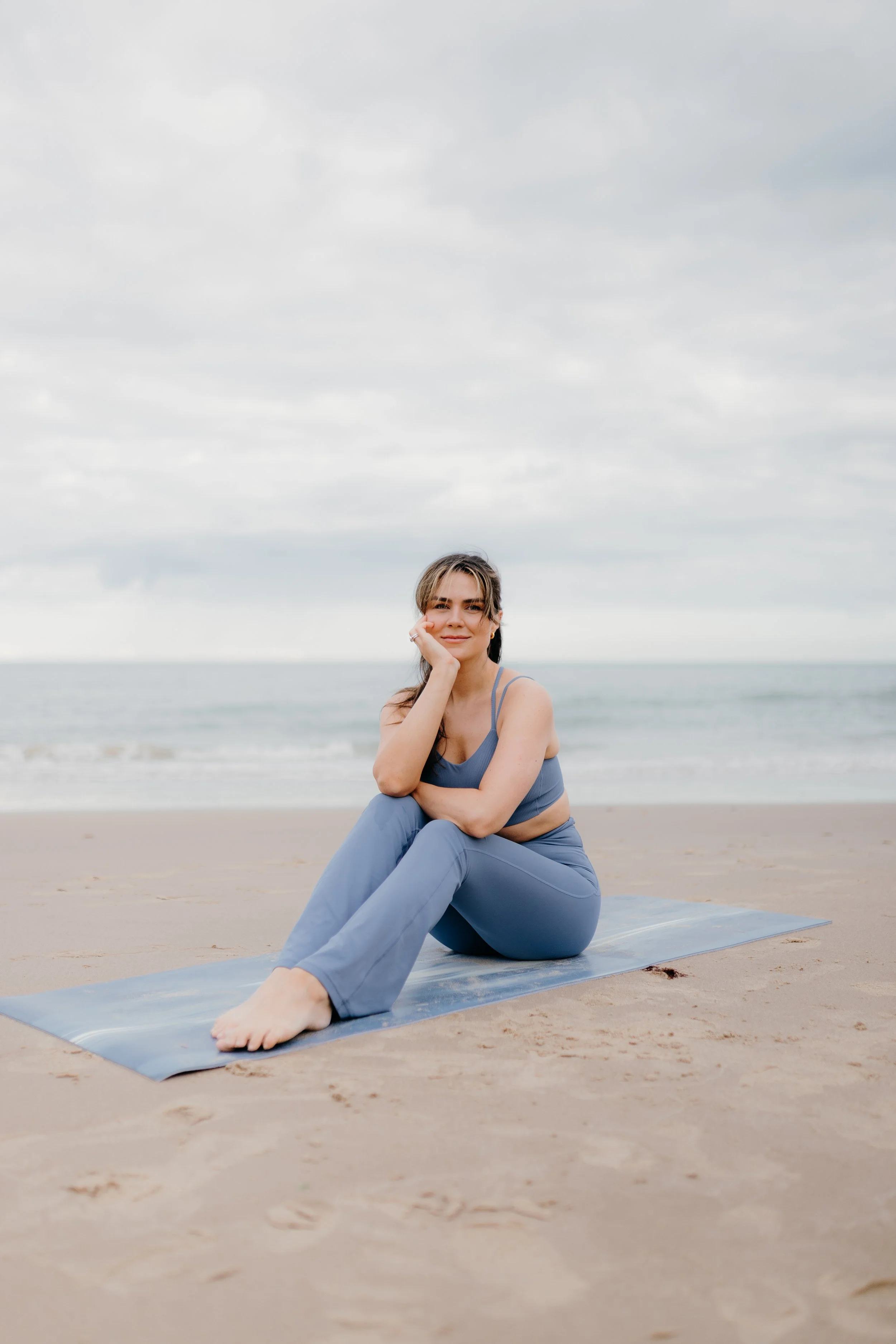 Woman sitting on yoga mat on the beach, smiling, with cloudy sky and ocean in the background.