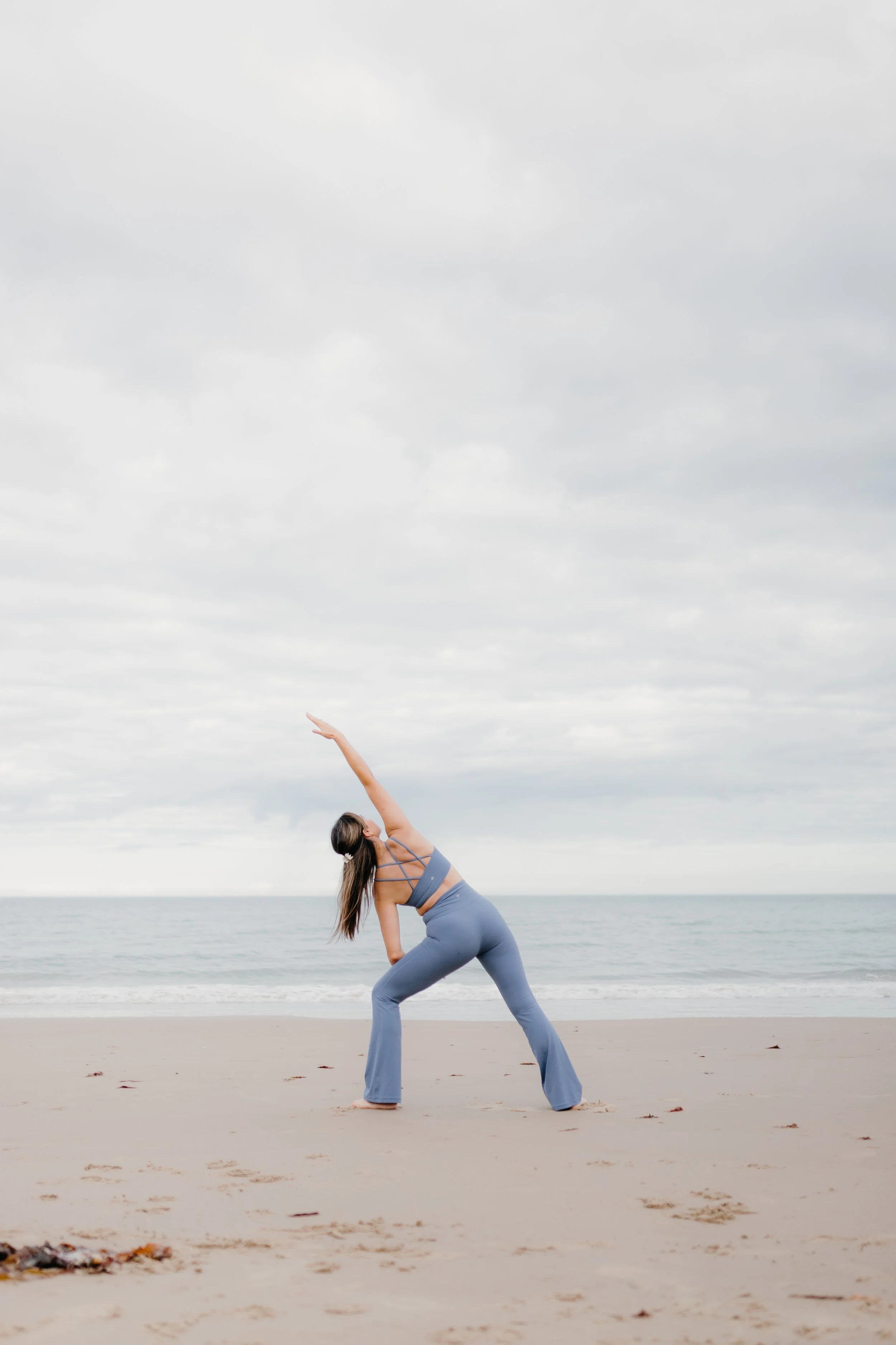 A woman practicing yoga on a beach, doing a side bend pose with one arm reaching overhead, wearing gray athletic wear, under cloudy skies, with the ocean in the background.