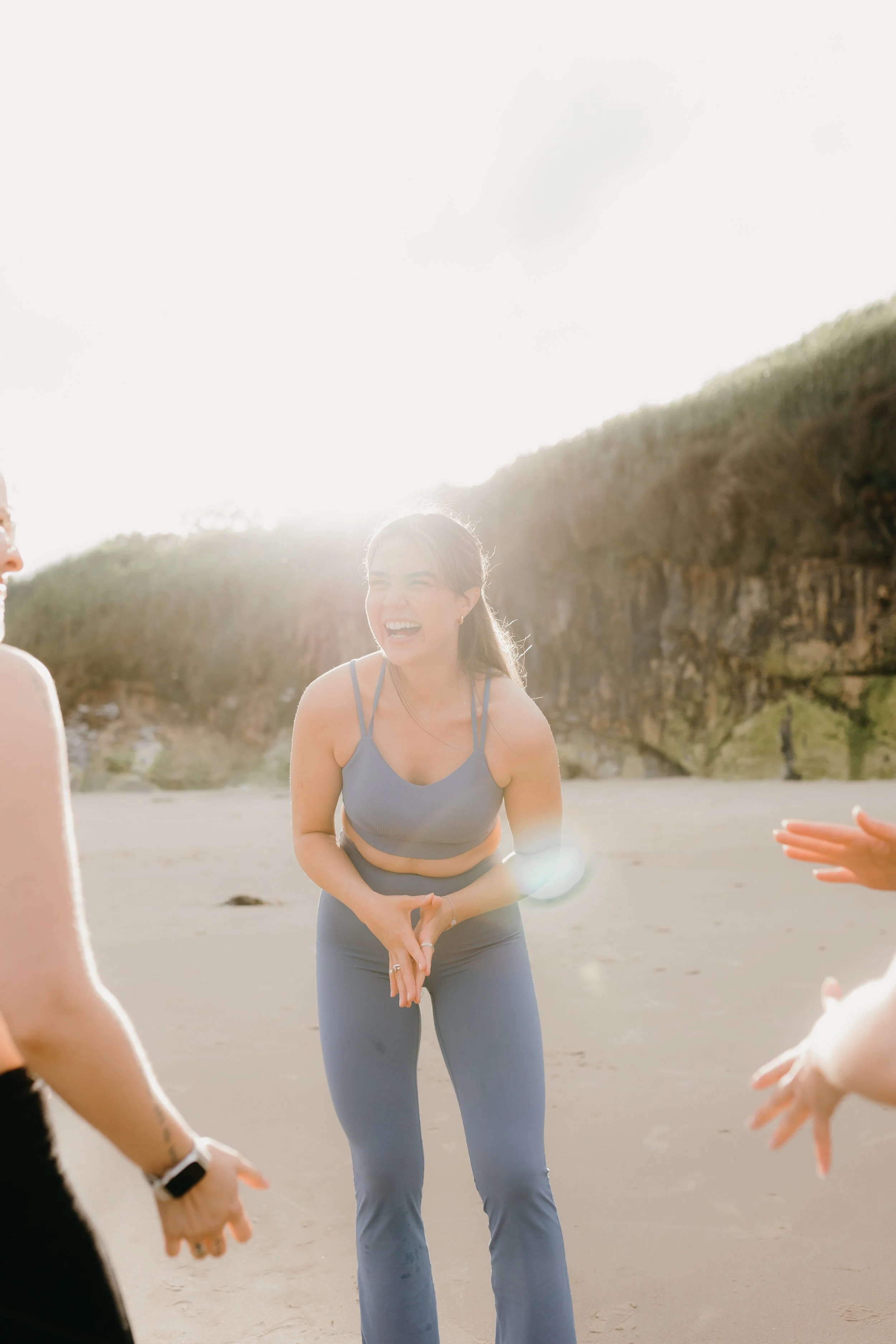 A young woman in athletic clothing smiling and laughing while standing outdoors on a sunny beach with cliffs in the background, surrounded by a group of people.