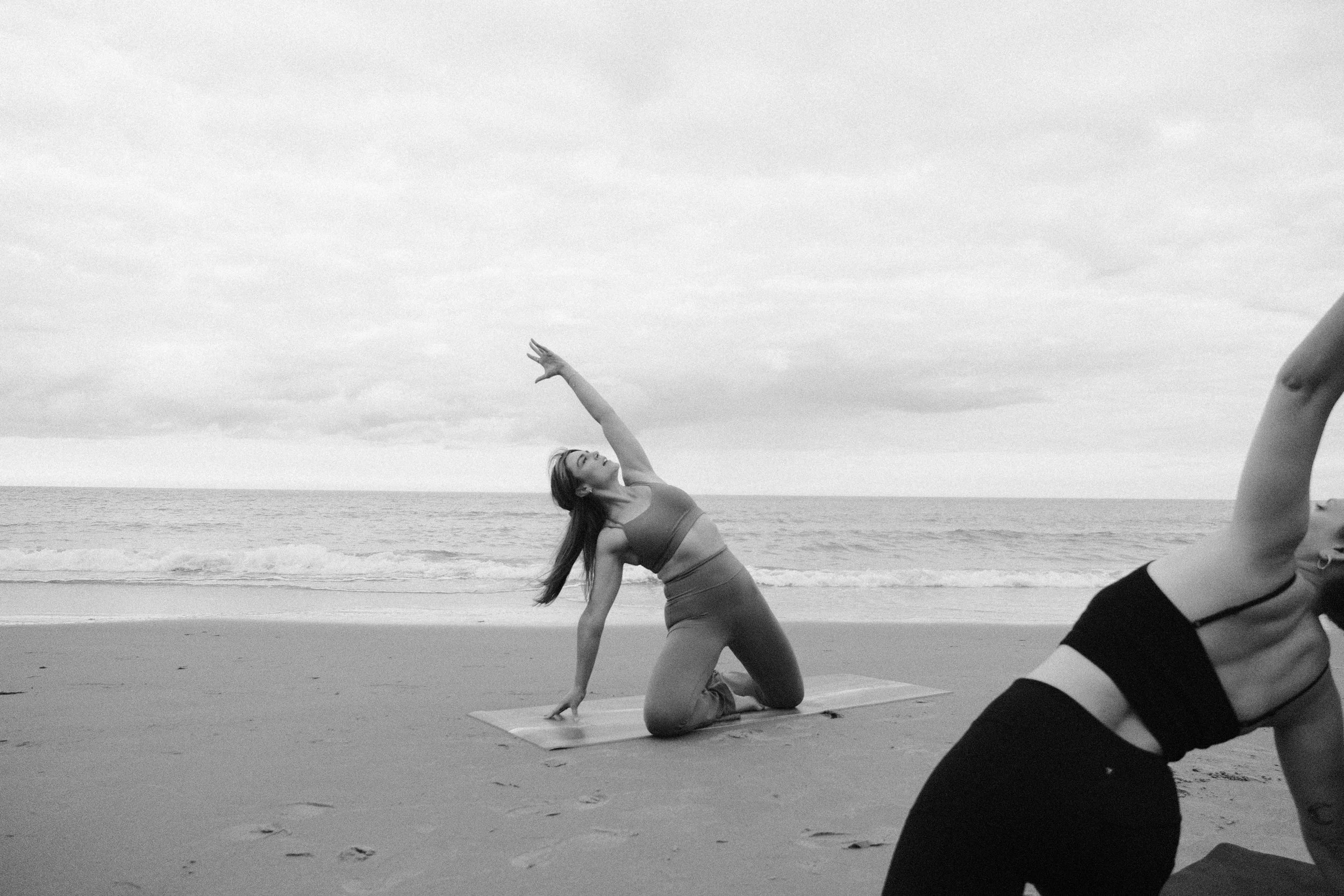 A woman practicing yoga on the beach, performing a side stretch pose on a yoga mat, with the ocean and cloudy sky in the background.
