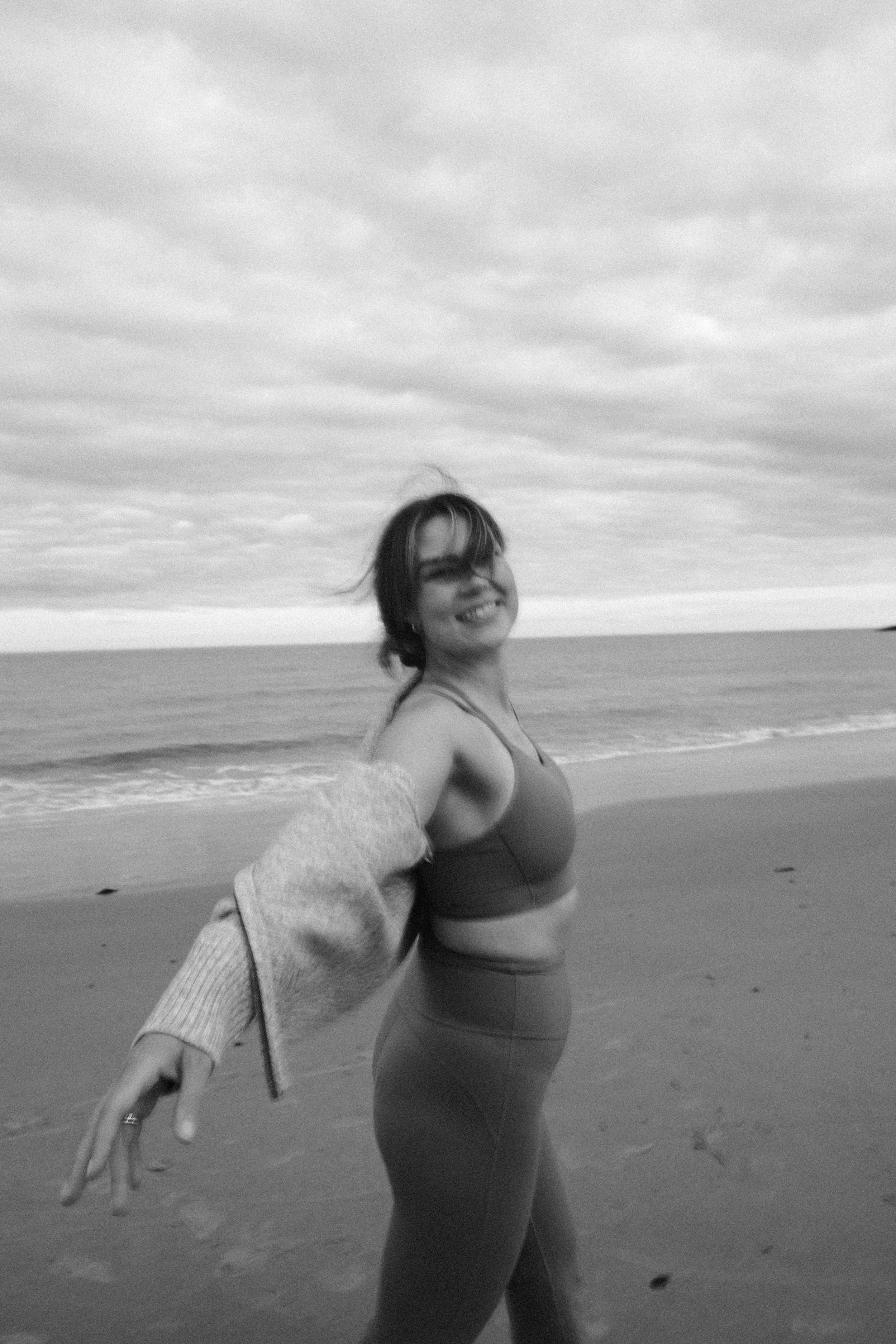 A woman smiling on a beach with her hand outstretched taking a selfie. She is wearing workout clothes, and the ocean and cloudy sky are in the background.