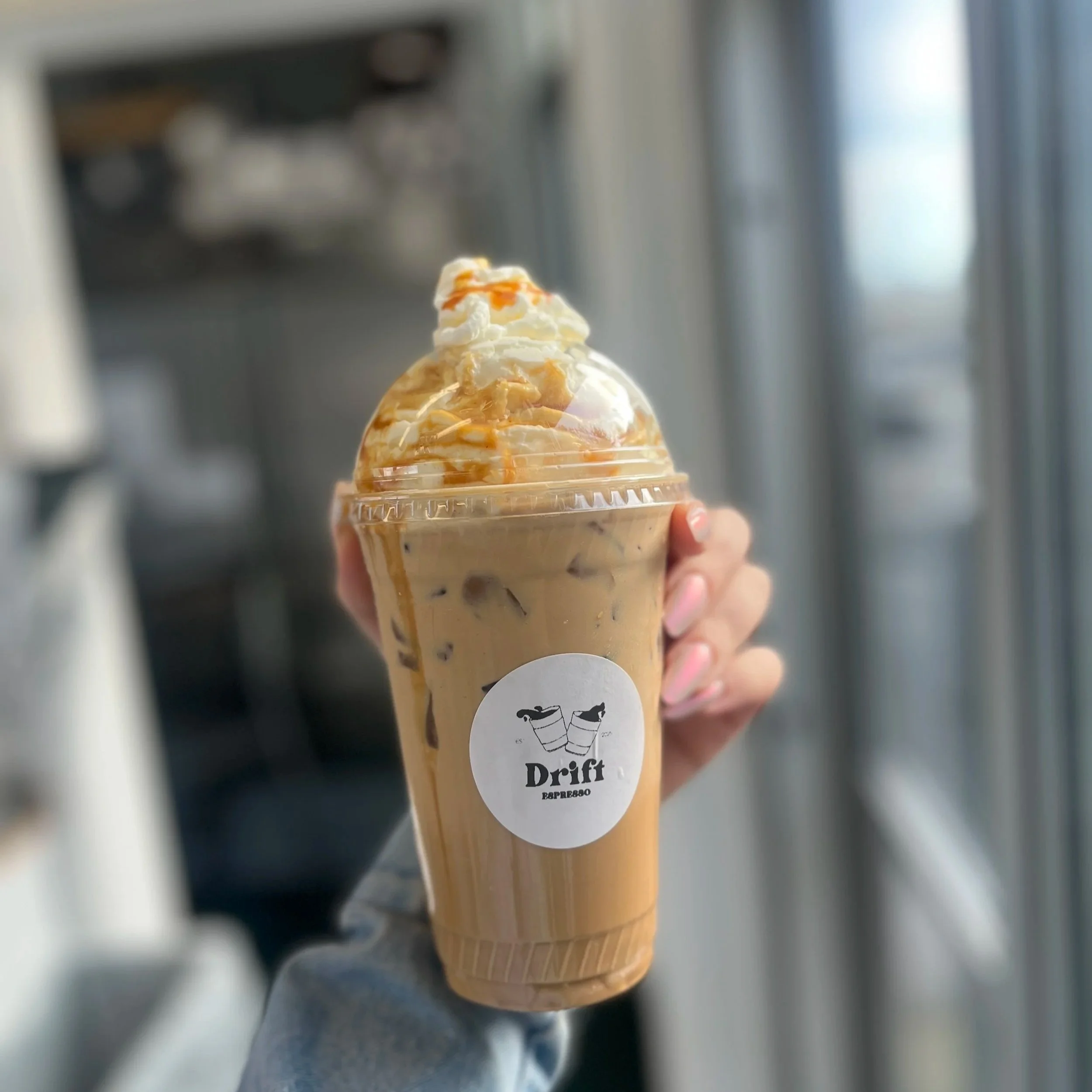 Close-up of hand holding an iced latte ice topped with whipped cream and caramel sauce, with a blurred background.