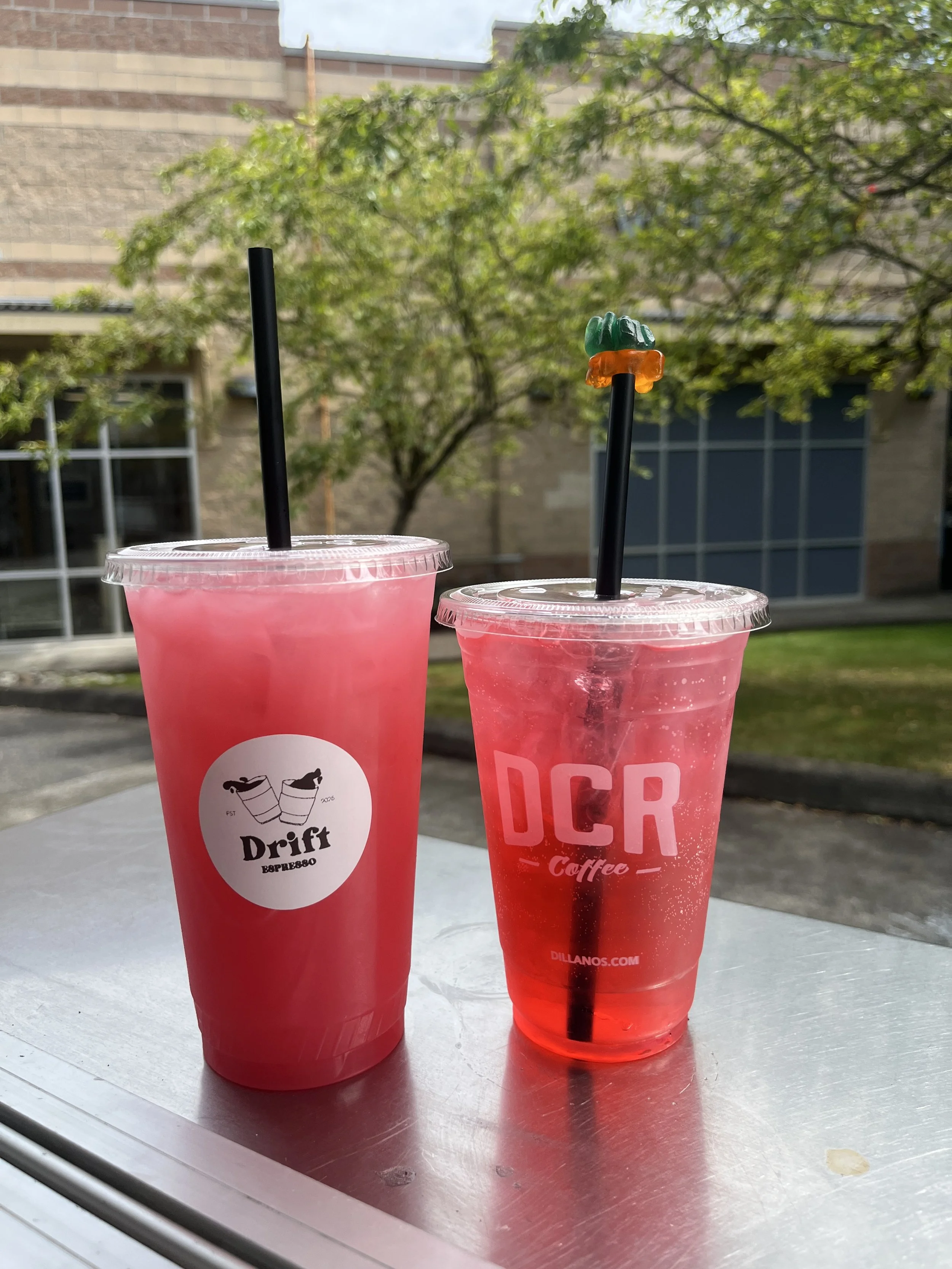 Two pink beverages in plastic cups with black straws, one cup has the drift espresso logo on it and the other has the word 'DCR' printed on it, placed on a metallic surface outdoors with trees and a building in the background.