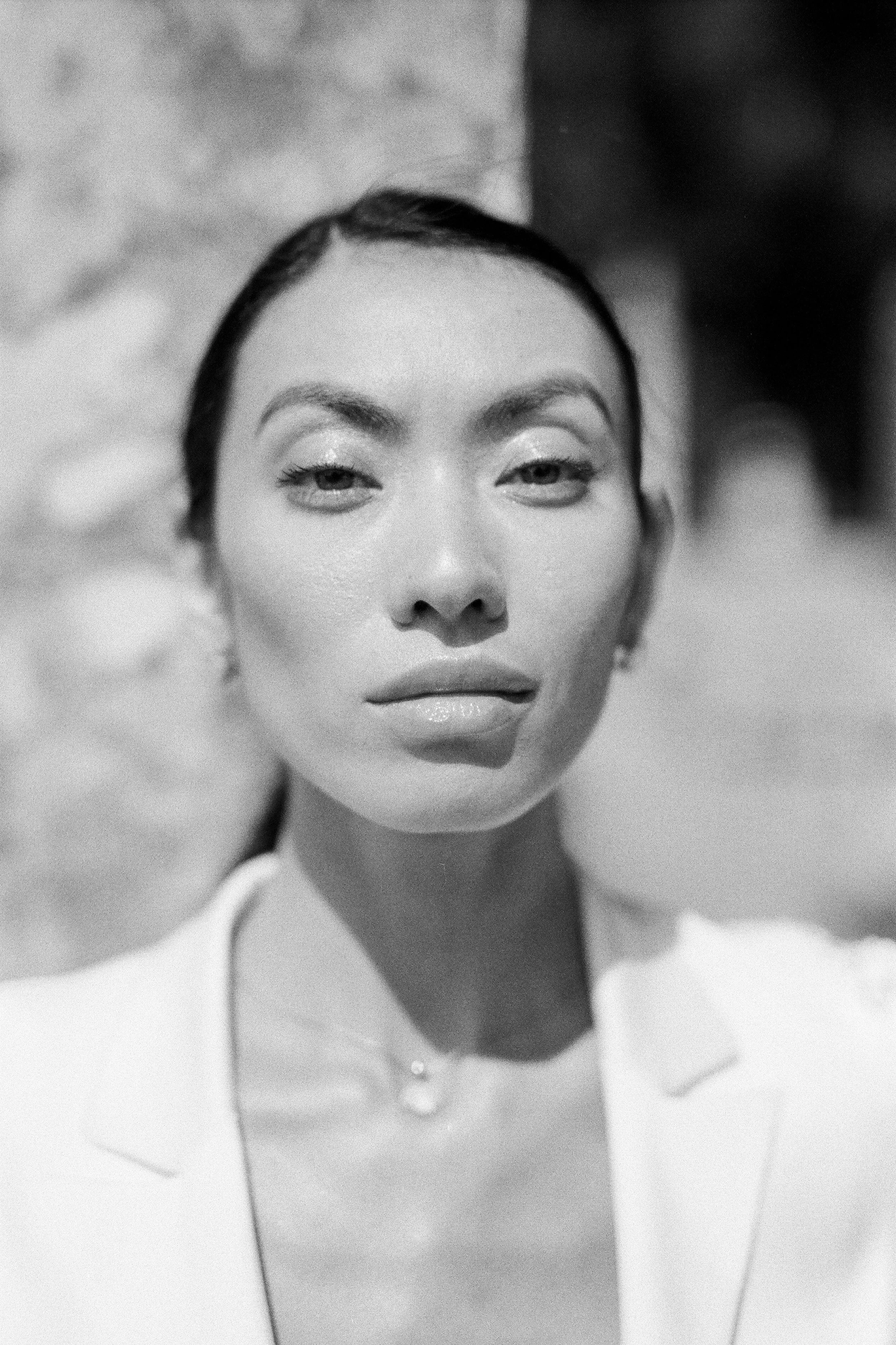 Black and white photo of a woman with makeup, wearing a white blazer and a necklace, looking at the camera.
