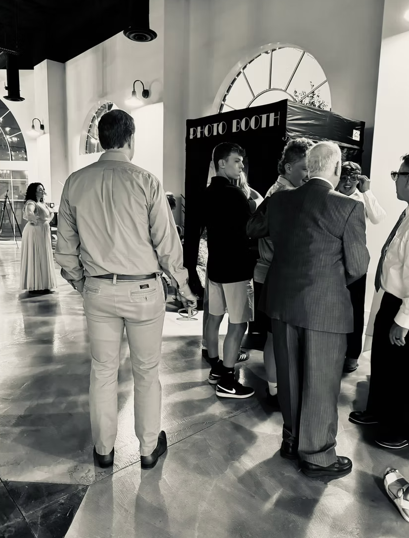 People gathered around a photo booth at an indoor event. Some are talking, and a woman is adjusting her goggles. Richly dressed individuals and casual attendees are present, with large windows in the background.