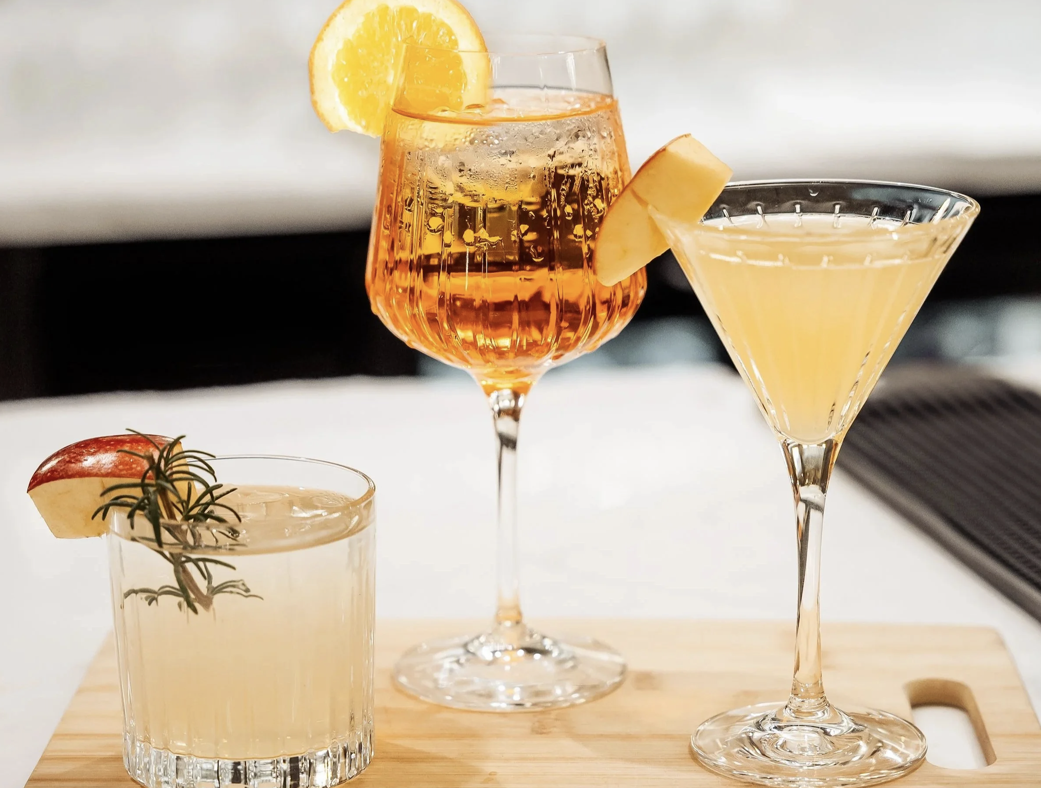 Three cocktails in tall glassware with lemon slices, set on a light wooden board, alongside a small tomato and rosemary garnish on a different glass.