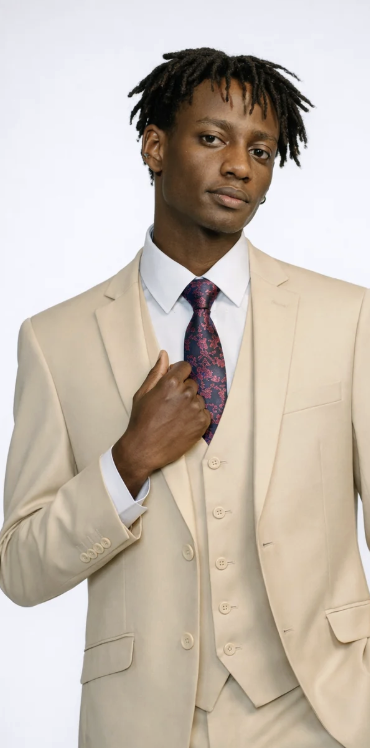 A young man with dreadlocks is dressed in a beige suit, white shirt, and a patterned tie, standing against a plain white background.