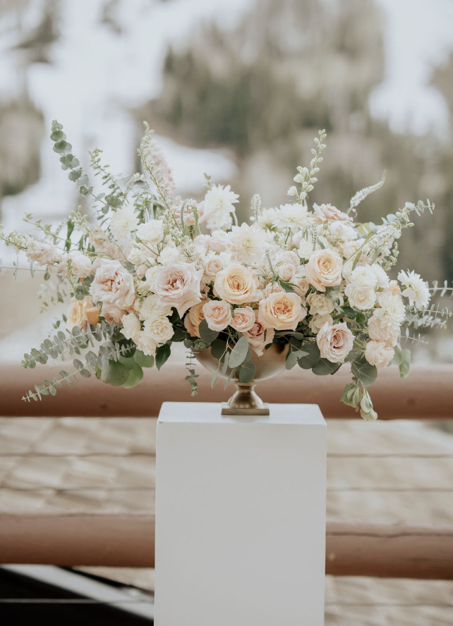 A floral arrangement with pale pink roses, white flowers, and green foliage in a decorative urn on a white pedestal.