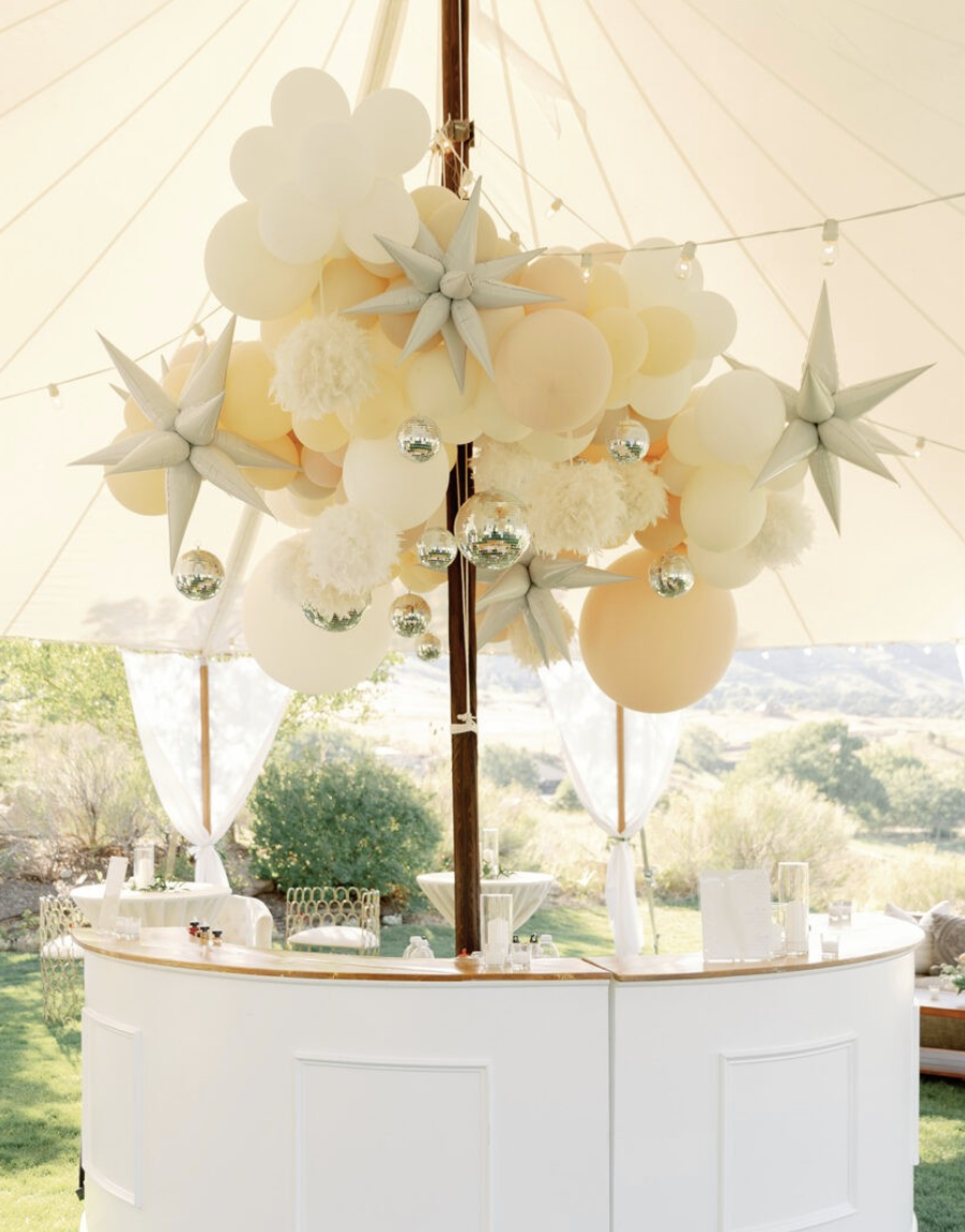 Decorative hanging arrangement of white spheres and metallic ornaments under a large cream-colored patio umbrella at outdoor event.