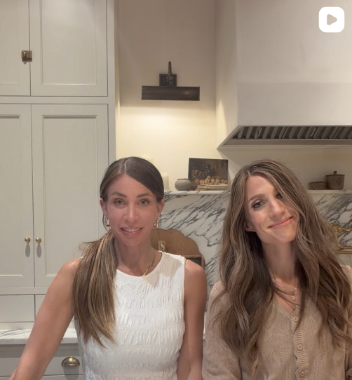 Two women sitting in a kitchen, smiling at the camera. The kitchen has white cabinets and a marble backsplash.