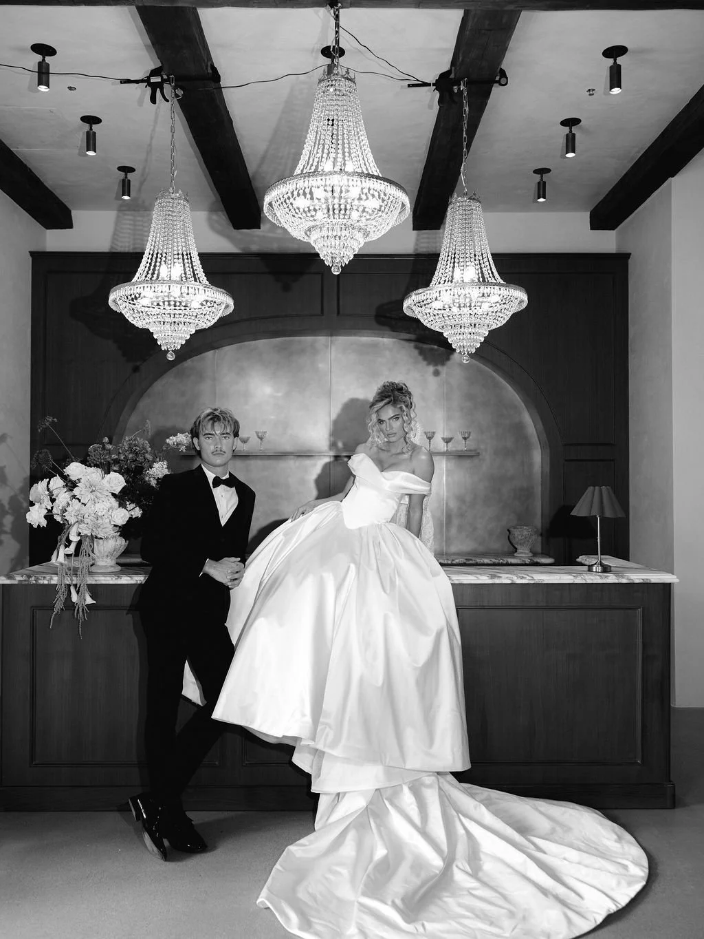 A black-and-white photo of a bride in a large wedding gown and a groom in a tuxedo, standing in a decorated room with chandeliers hanging from the ceiling.
