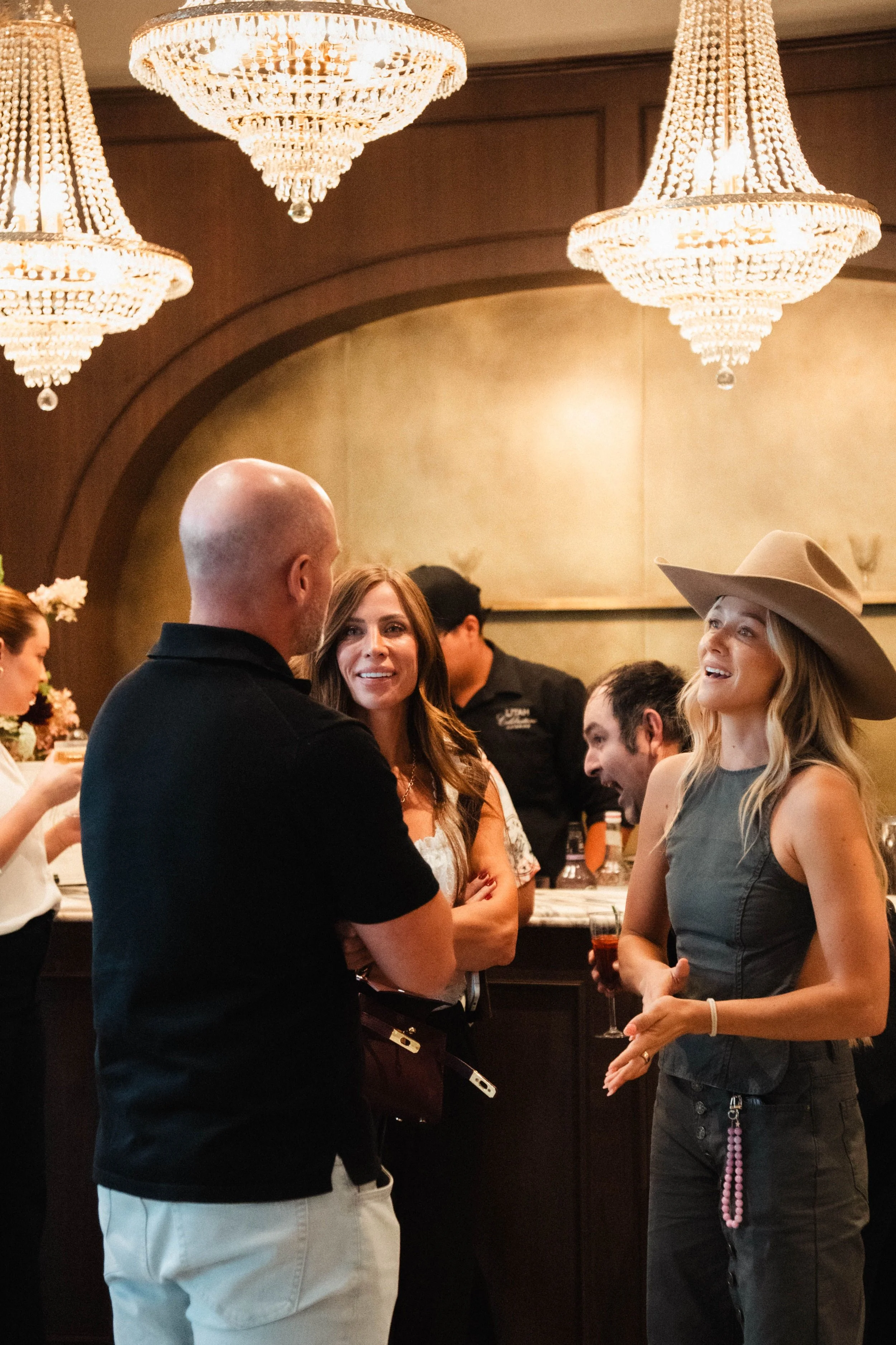 People socializing at a bar or restaurant, with crystal chandeliers hanging from the ceiling, some holding drinks, and a woman wearing a wide-brimmed hat and necklace of pink beads.