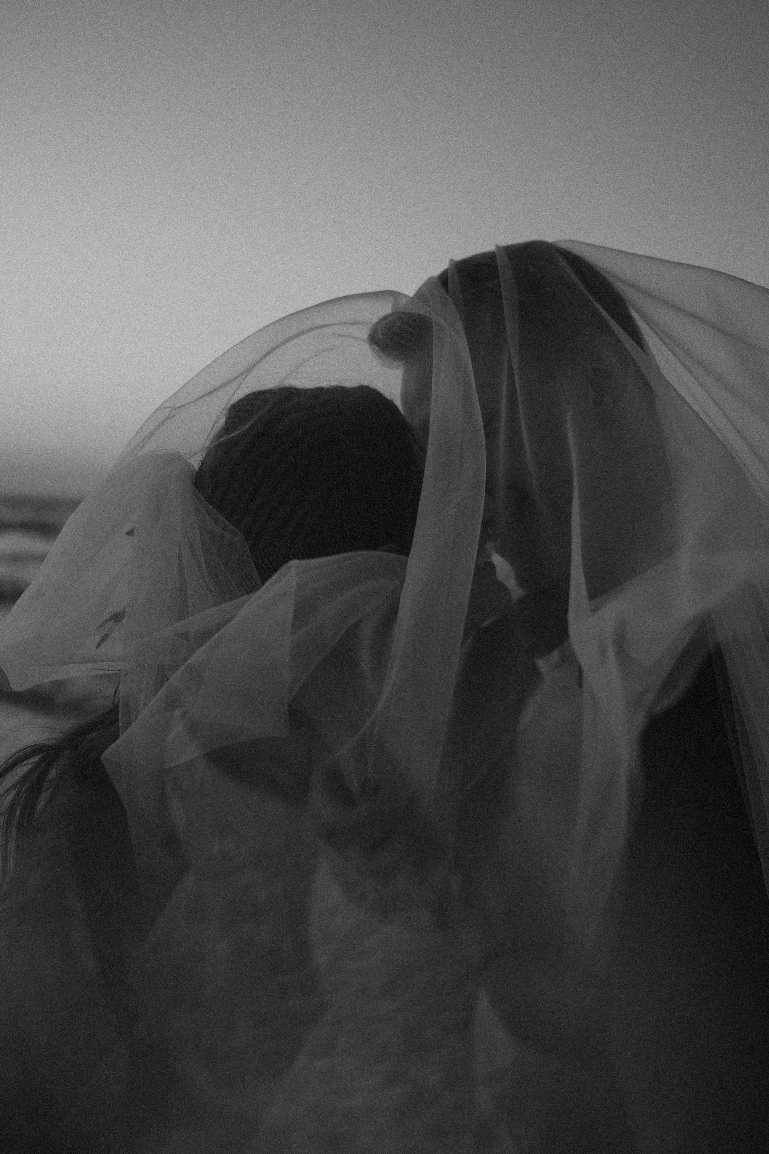 Black and white photo of a person wearing a face mask and a transparent veil, with the sky in the background.