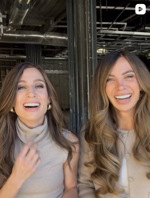 Two women with long, wavy hair smiling and laughing, sitting indoors with visible metal pipes and beams overhead.