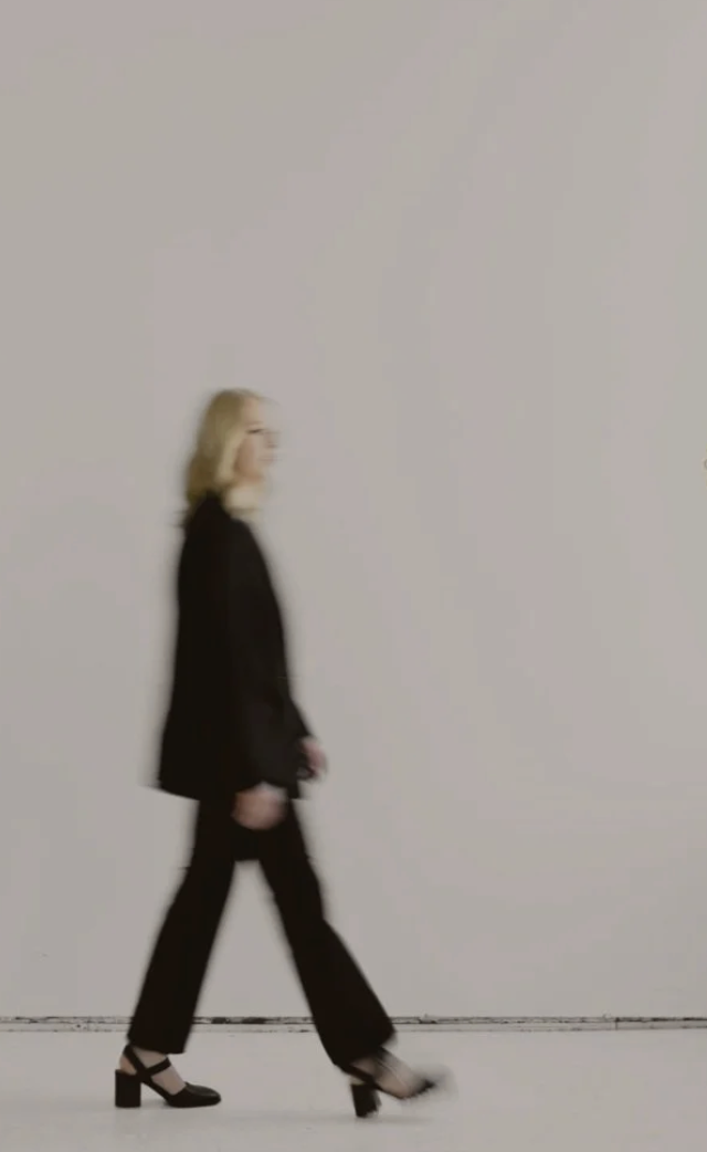 Woman with blonde hair walking past a plain light-colored wall, wearing a black blazer, black pants, and black high heels.