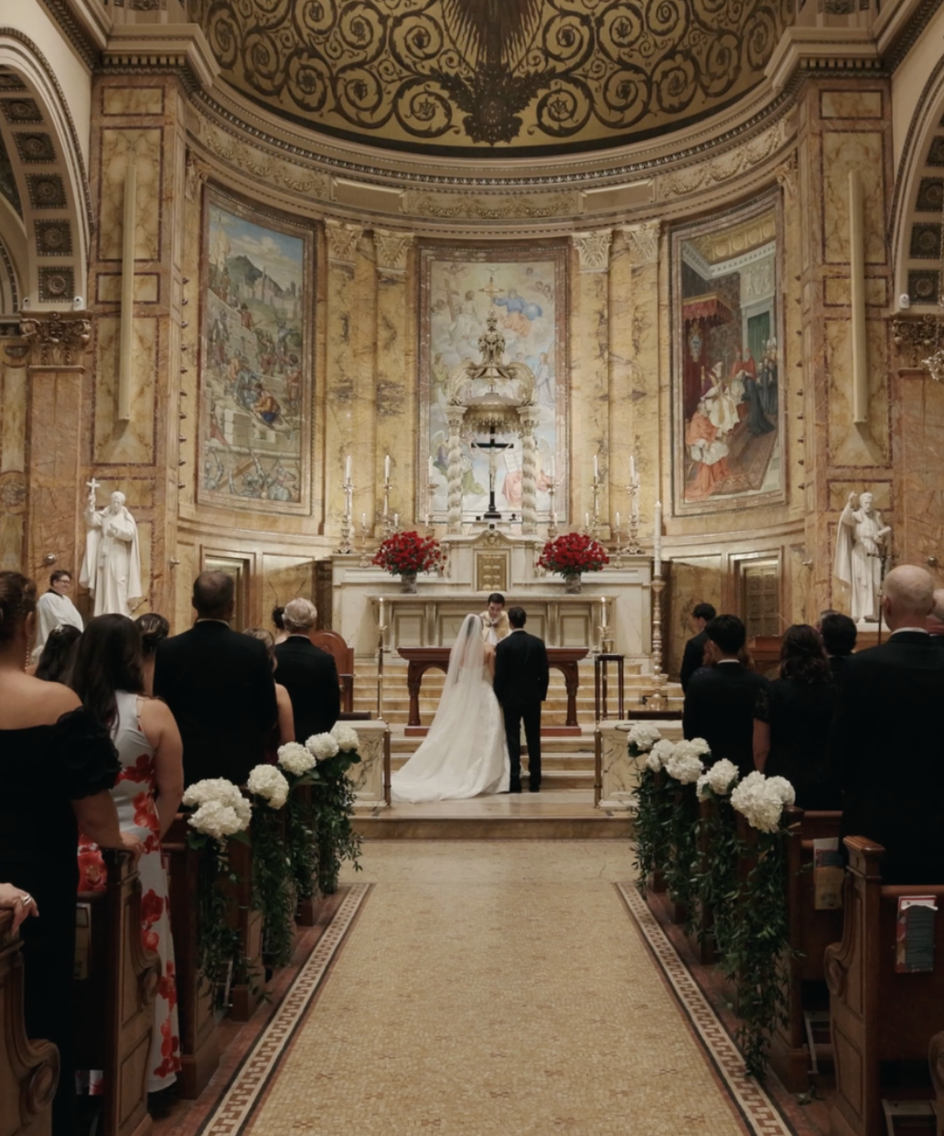 A wedding ceremony taking place in a grand church with ornate gold and marble interior, stained glass murals, and statues. The bride and groom stand at the altar in front of a priest, with guests seated on either side.