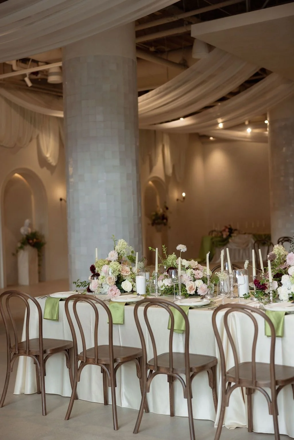 Elegant banquet table decorated with pastel-colored flowers and candles in a decorated event space with draped ceilings and columns.