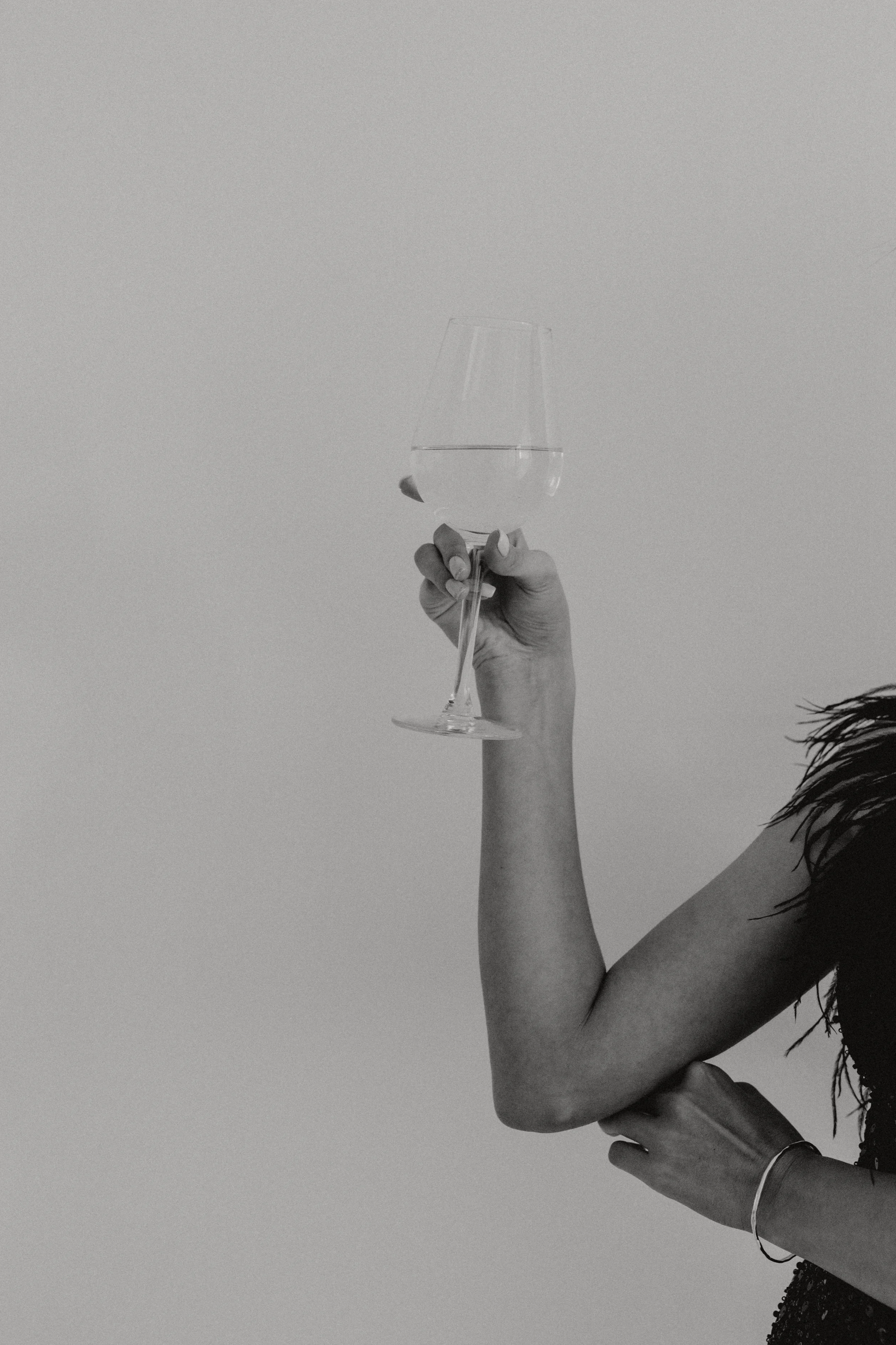 A person holding a wine glass filled with water against a plain background. The person's arm is visible, with a bracelet, and part of their body is shown, dressed in a dark, sparkly top.