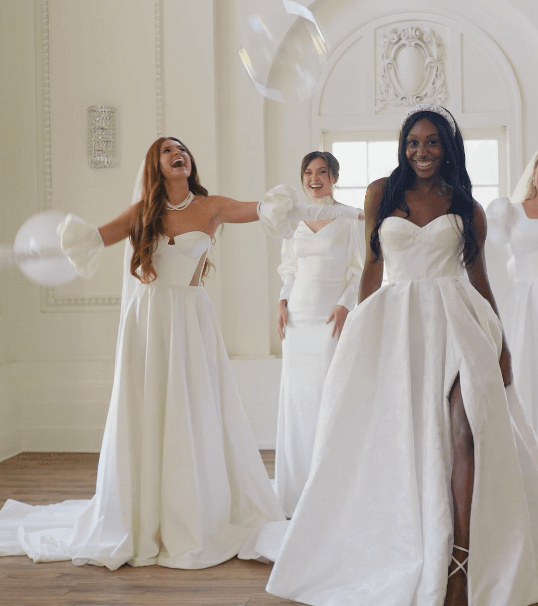 Women in wedding dresses celebrating in a bright, elegant room with high ceilings and large windows, surrounded by floating balloons.