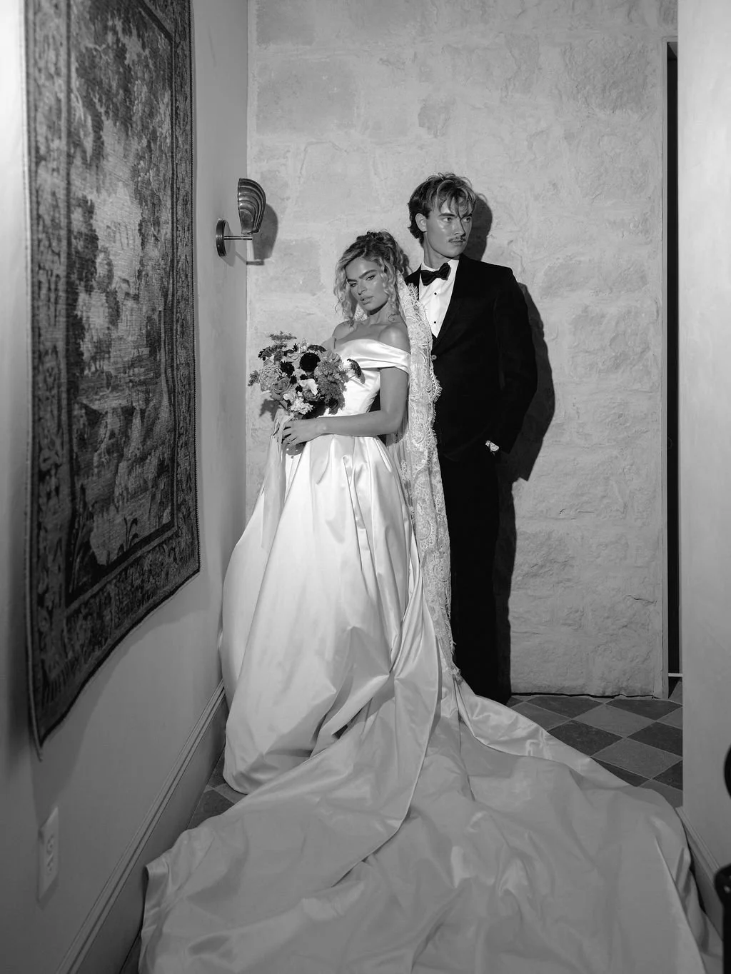 Black and white image of a bride and groom standing in a hallway. The bride is holding a bouquet, wearing an off-the-shoulder wedding gown and veil, with a serious expression. The groom is dressed in a tuxedo, standing behind her with his hands in his pockets, looking away. There is a textured wall and a carpeted floor.