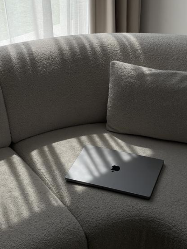 A closed silver MacBook laptop resting on a light beige fabric couch with sunlight streaming through a window, creating shadows.