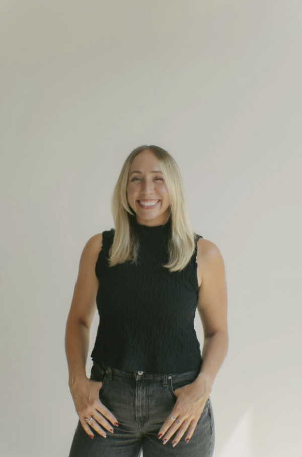 A young woman with shoulder-length blonde hair, smiling, wearing a sleeveless black top and dark jeans, standing against a plain light-colored wall.