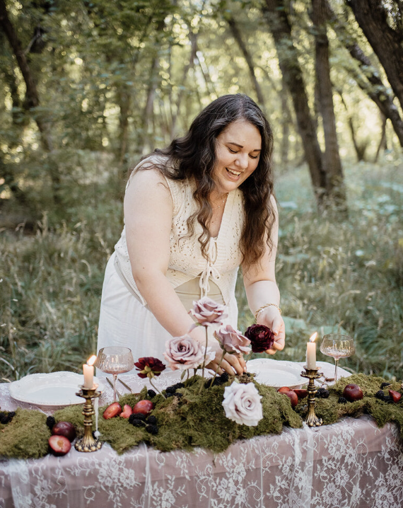 A woman with long dark hair wearing a light-colored dress setting a table outdoors in a wooded area, surrounded by flowers, candles, glasses, and apples.