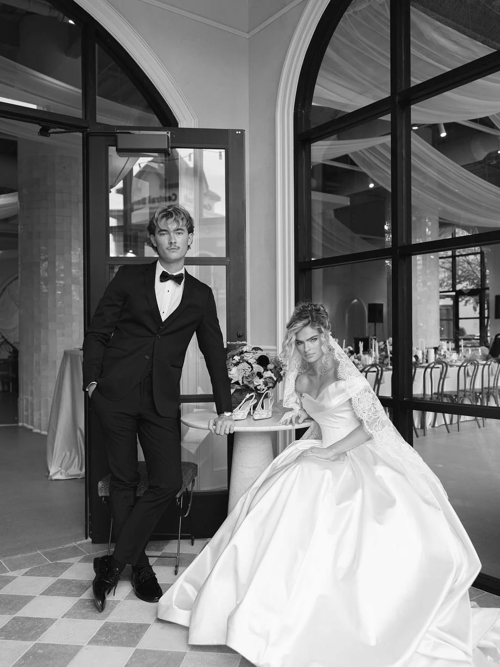 A black and white photo of a young couple in wedding attire inside a well-lit venue, with large arched windows and interior tables. The groom, dressed in a tuxedo, stands with his hand in his pocket next to the bride, who is seated at a small round table wearing a wedding gown and veil. There is a floral bouquet and a pair of high-heeled shoes on the table.