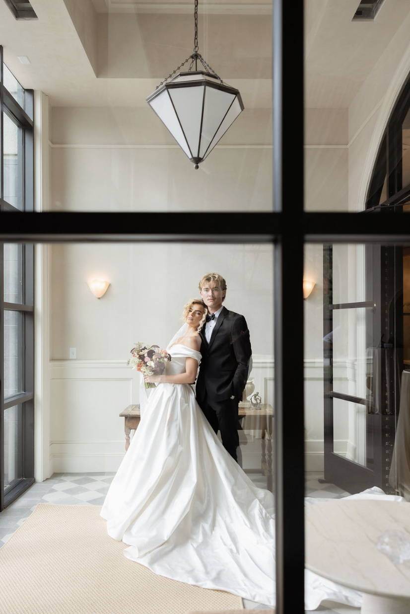 A bride and groom stand together in an elegant room, the bride holding a bouquet and wearing a white wedding gown, the groom in a black suit and bow tie, viewed through a window.