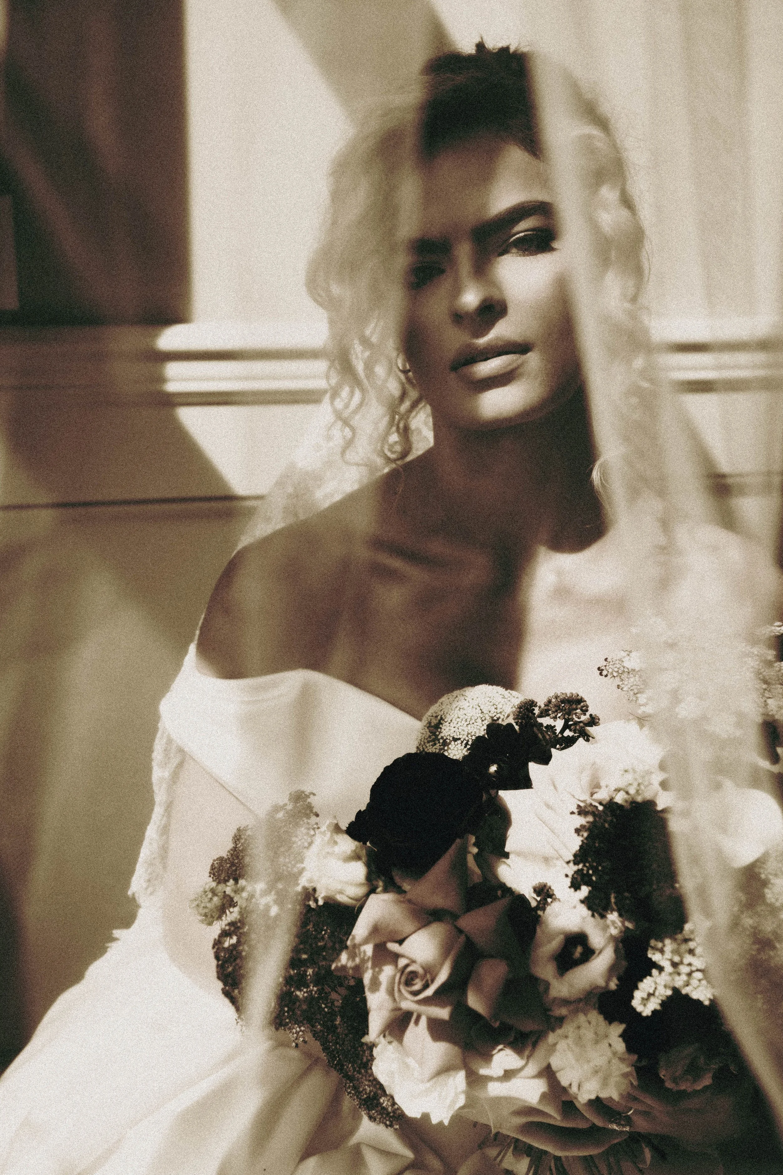 Black and white photograph of a woman with curly hair and elegant makeup, holding a bouquet of roses and flowers, wearing an off-shoulder dress, posing indoors near a window.